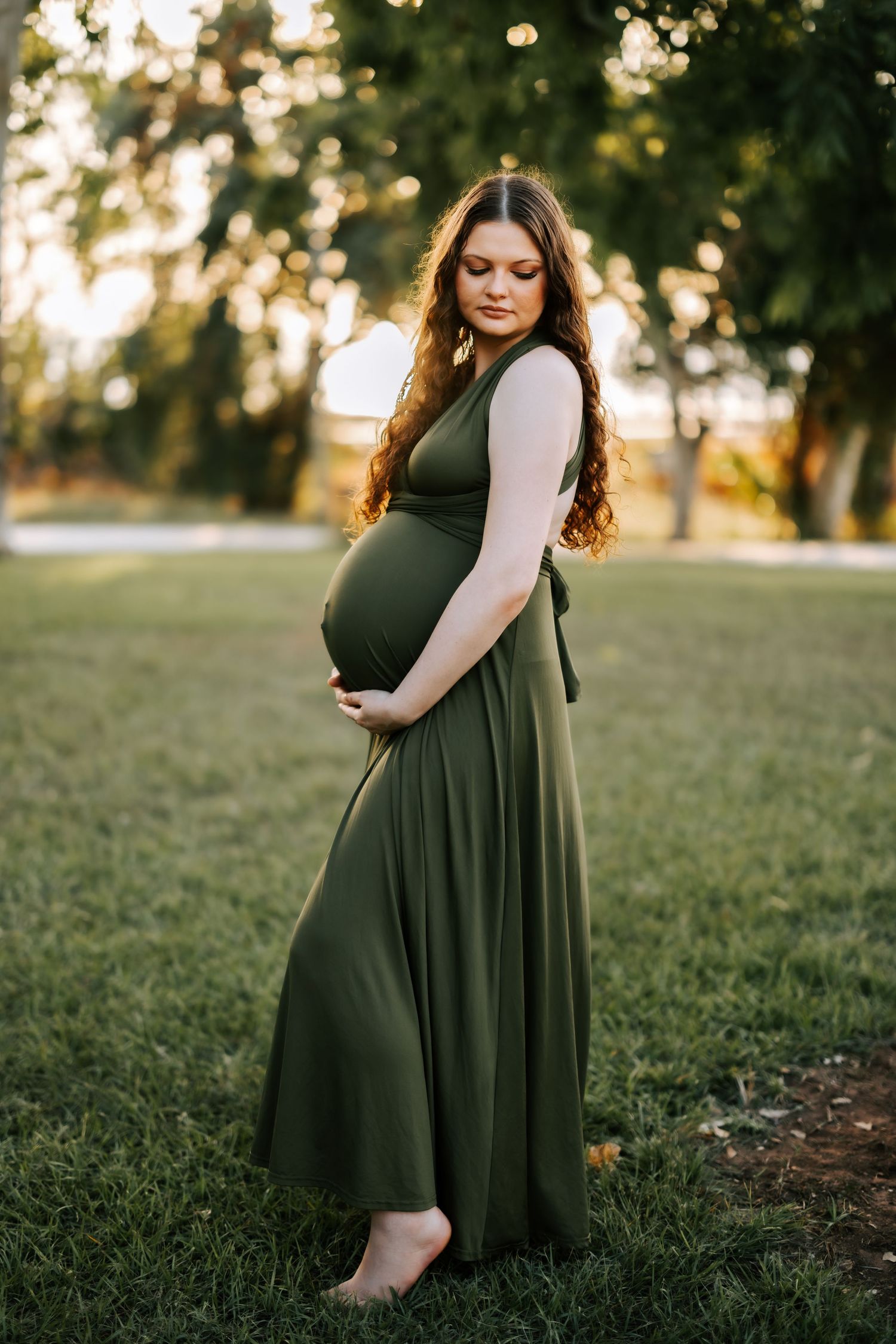 Pregnant woman in olive green maxi dress cradling baby bump in a sunlit park.