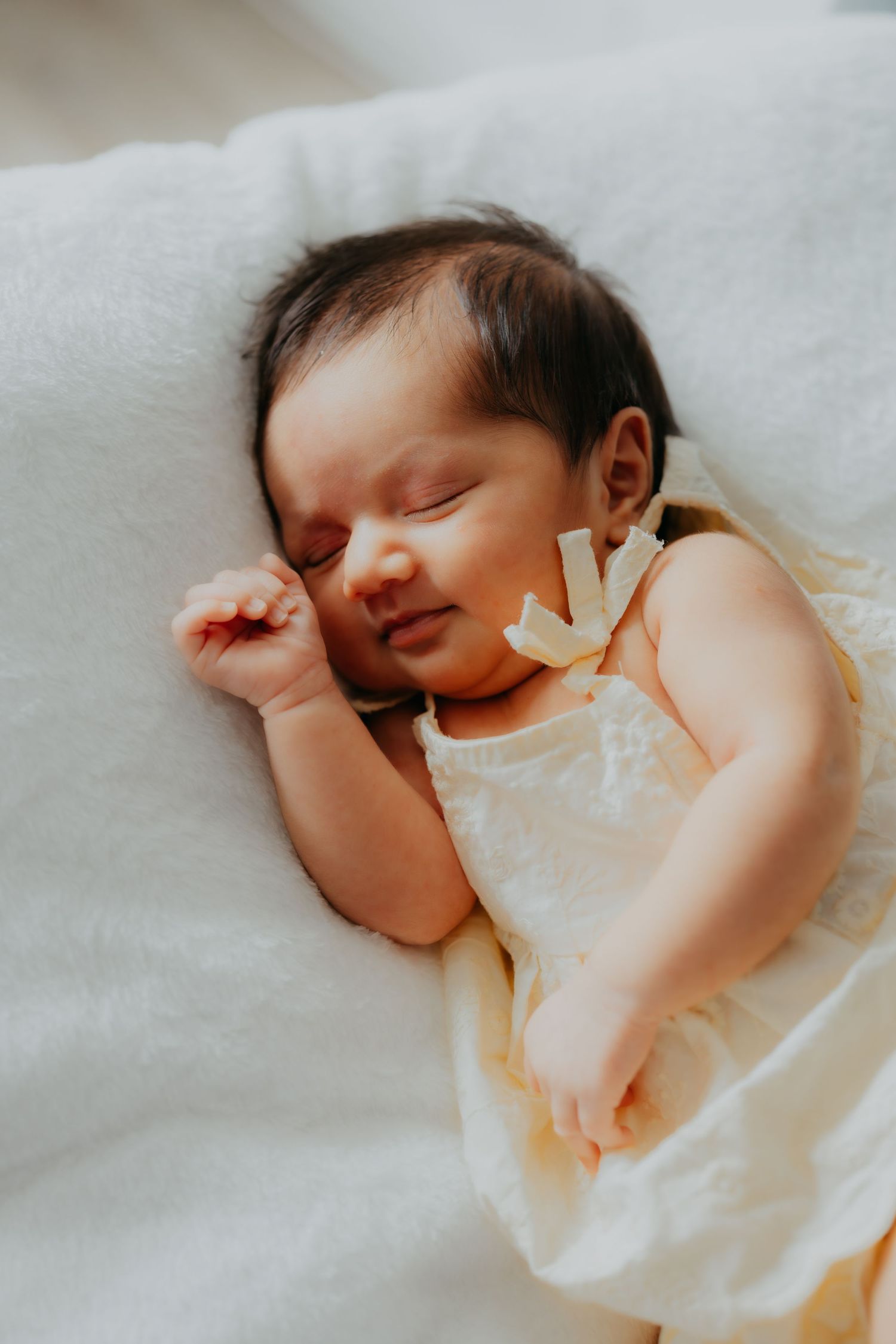 Peaceful newborn baby girl sleeping in a cream dress on a white fluffy blanket.