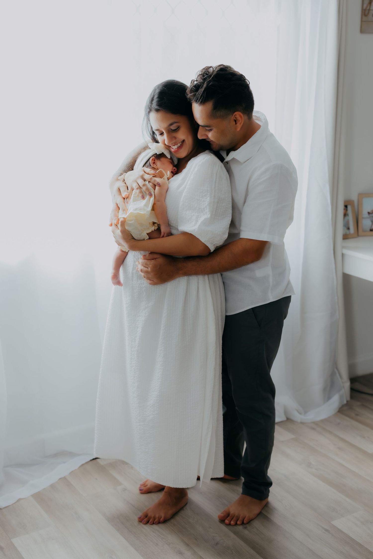 Smiling couple in white outfits holding their newborn baby in a bright airy room.