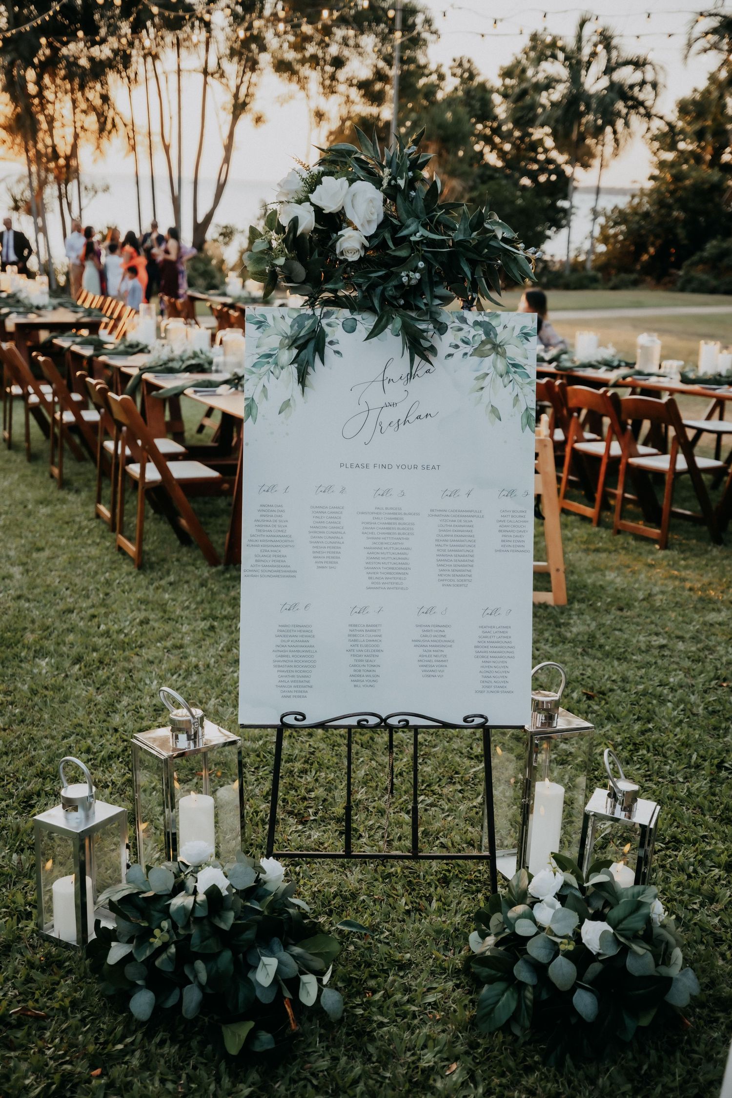 Elegant outdoor wedding seating chart display with white floral greenery decor and lanterns on grass.