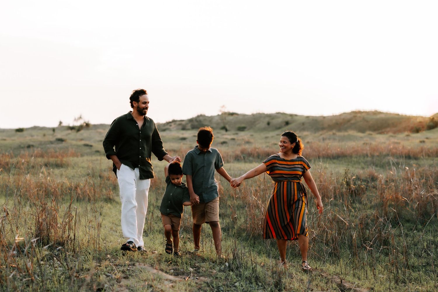 Family of four holding hands while walking through golden field at sunset outdoors.
