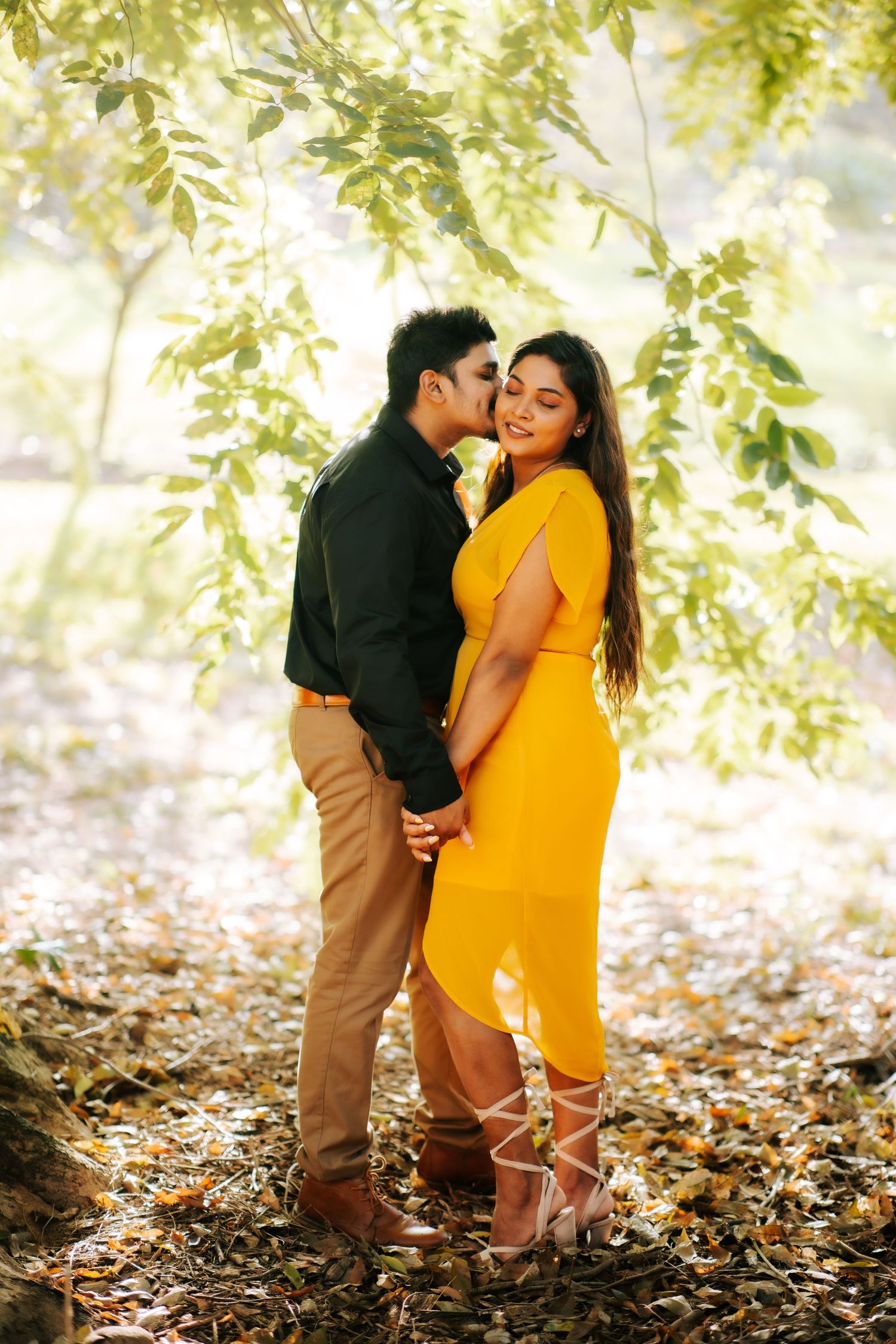 Couple embracing under sunlit green leaves, woman in yellow dress being kissed on cheek by partner.