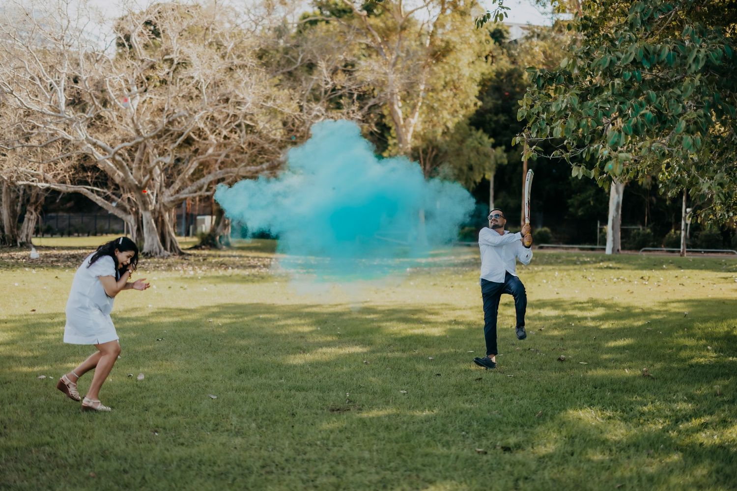 Couple doing a gender reveal with blue powder smoke in an outdoor park setting surrounded by trees.