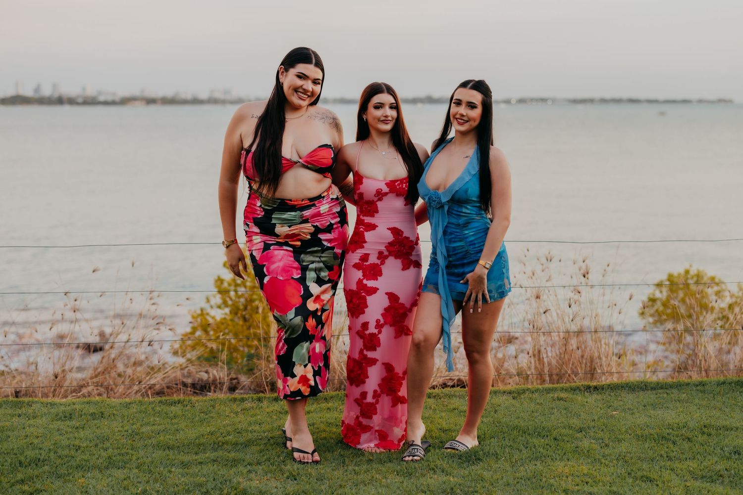 Three young women in colorful summer dresses posing together outdoors near a waterfront.