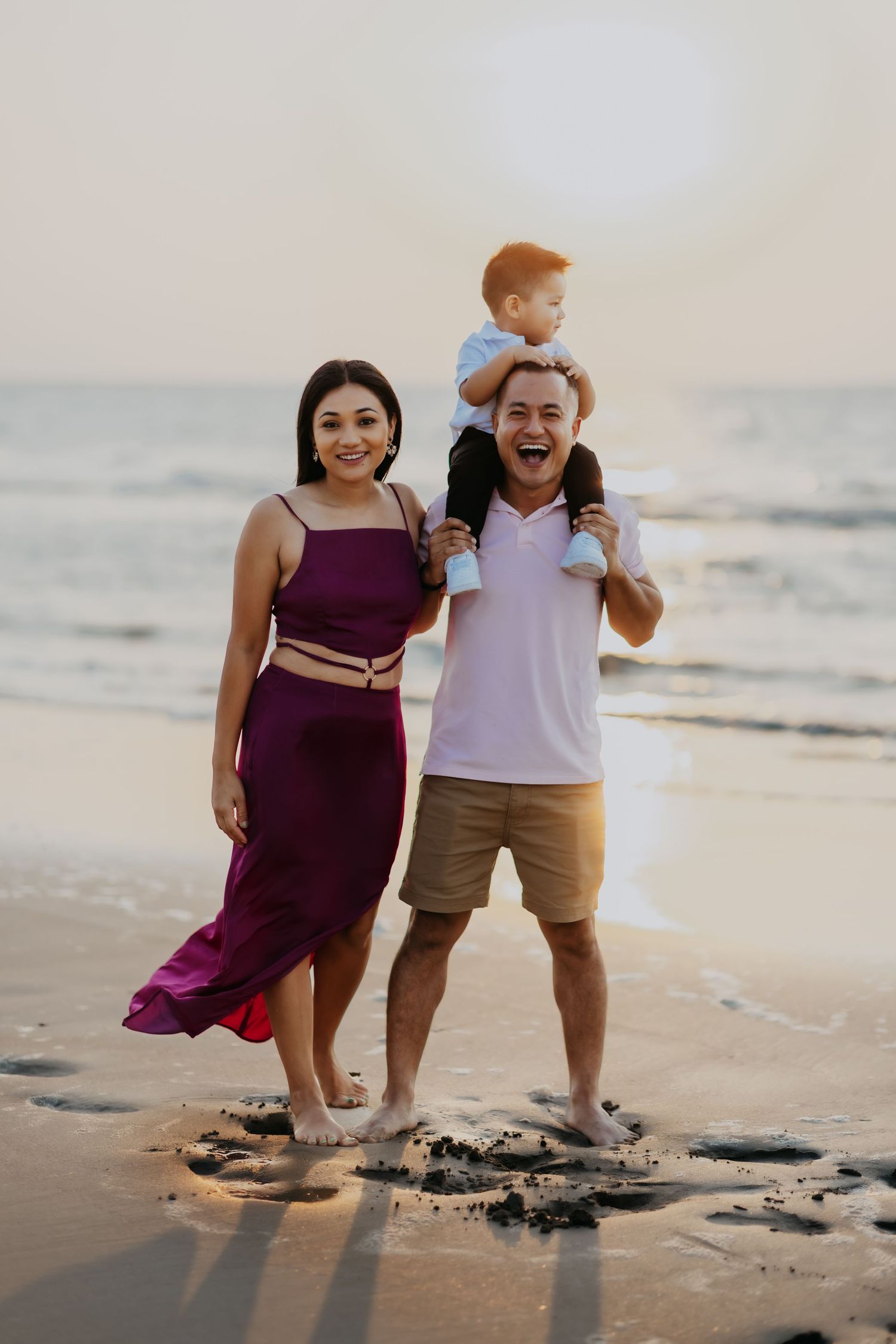 A smiling family poses on the beach at sunset, with dad carrying toddler son on shoulders.