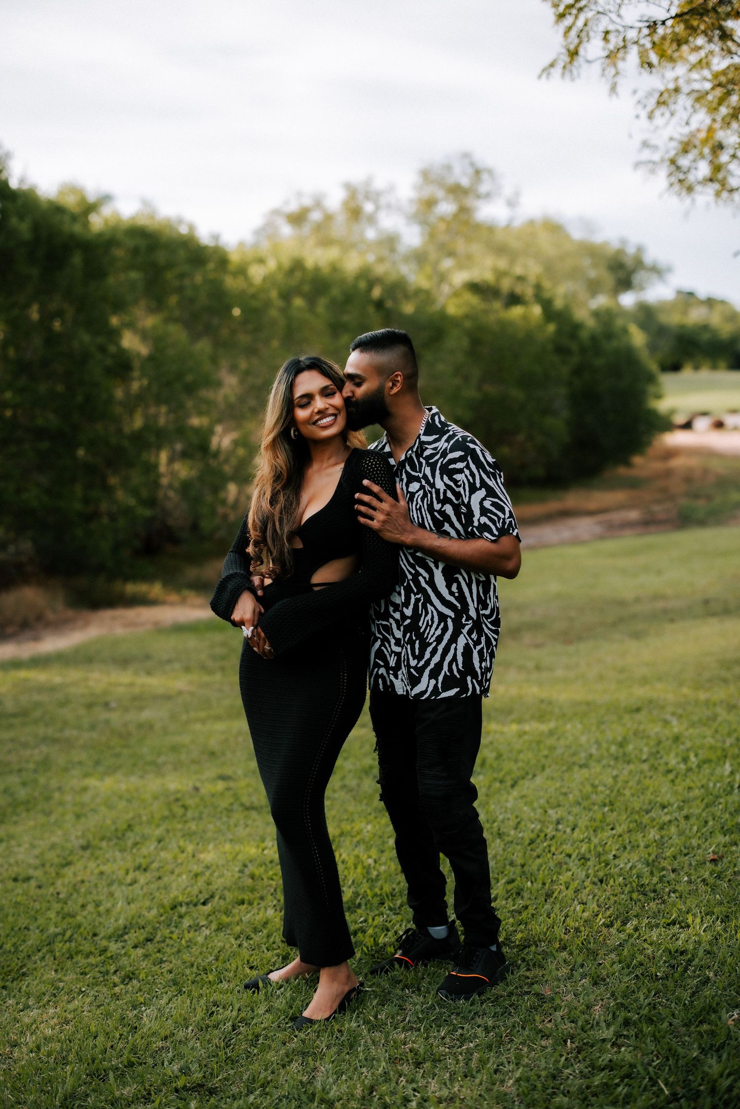 A couple embraces outdoors, woman in black outfit smiling as man in zebra print shirt kisses her cheek on green lawn.