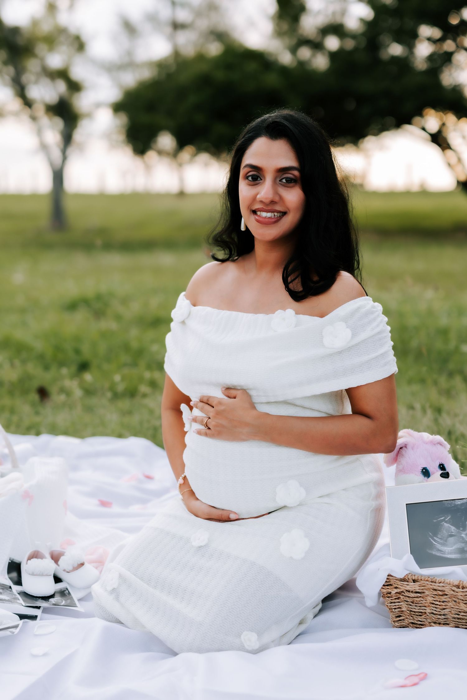Smiling pregnant woman in white off-shoulder dress sitting on a blanket in a park at golden hour.