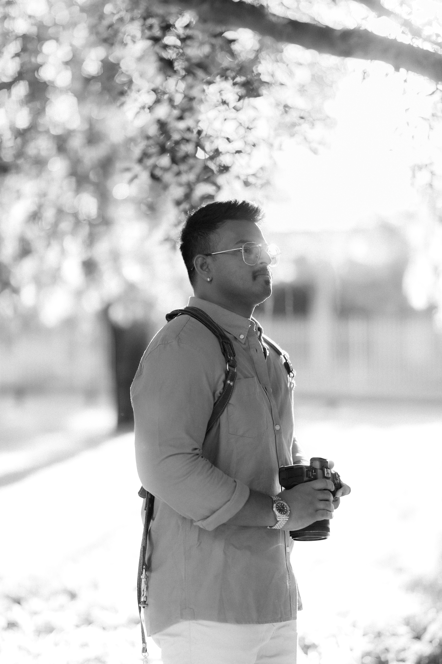 A man holding a camera stands under a tree in a black and white outdoor portrait.