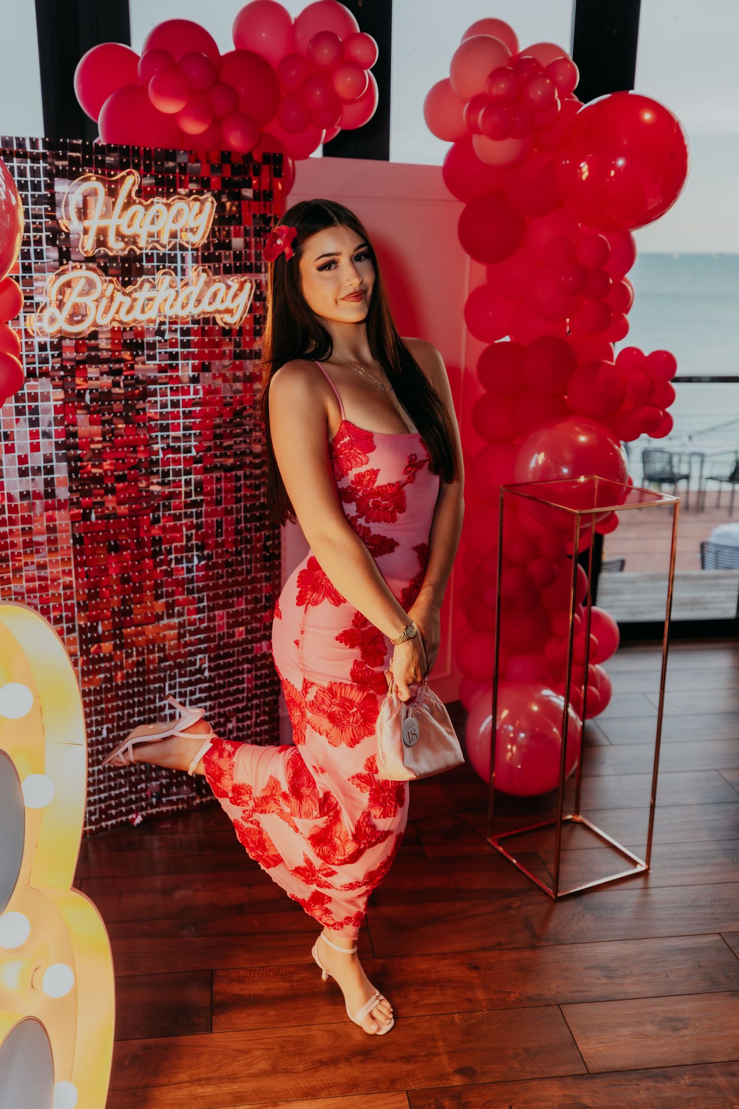 Woman in red floral dress poses at birthday party with red balloon arch and Happy Birthday sign.