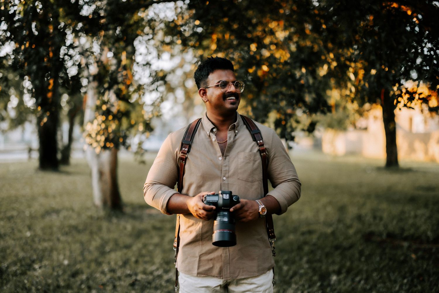 Smiling male photographer holding camera while standing in a sunlit park with trees.