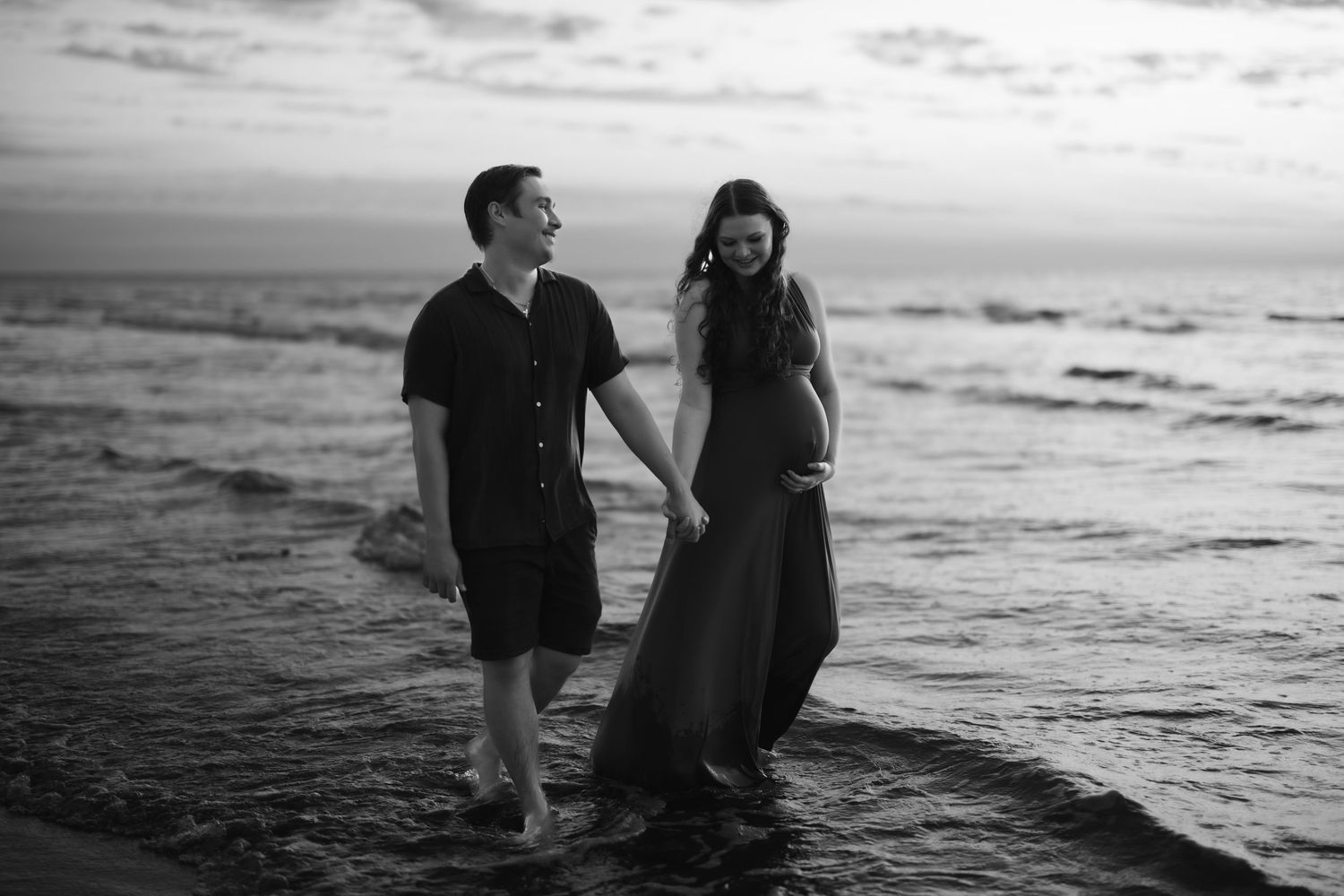 Black and white photo of a couple holding hands while standing in ocean waves at sunset on the beach.