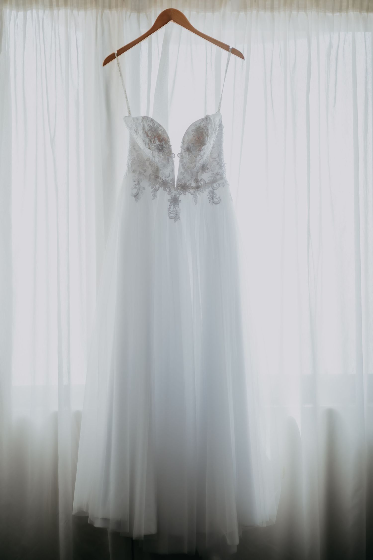 Elegant white wedding dress with lace bodice hanging on a wooden hanger by a window.
