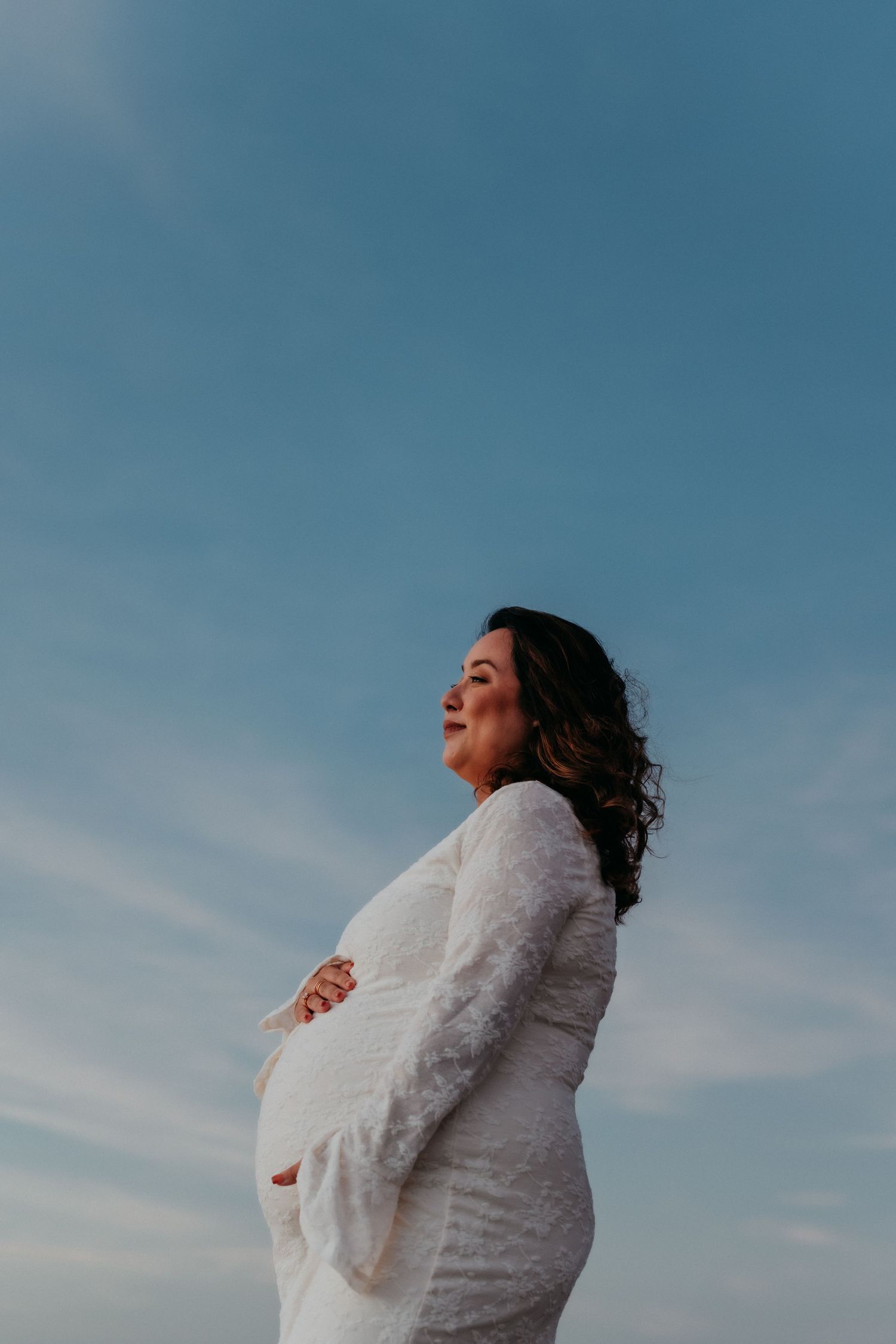 Pregnant woman in white lace dress holding her baby bump against a blue sky.