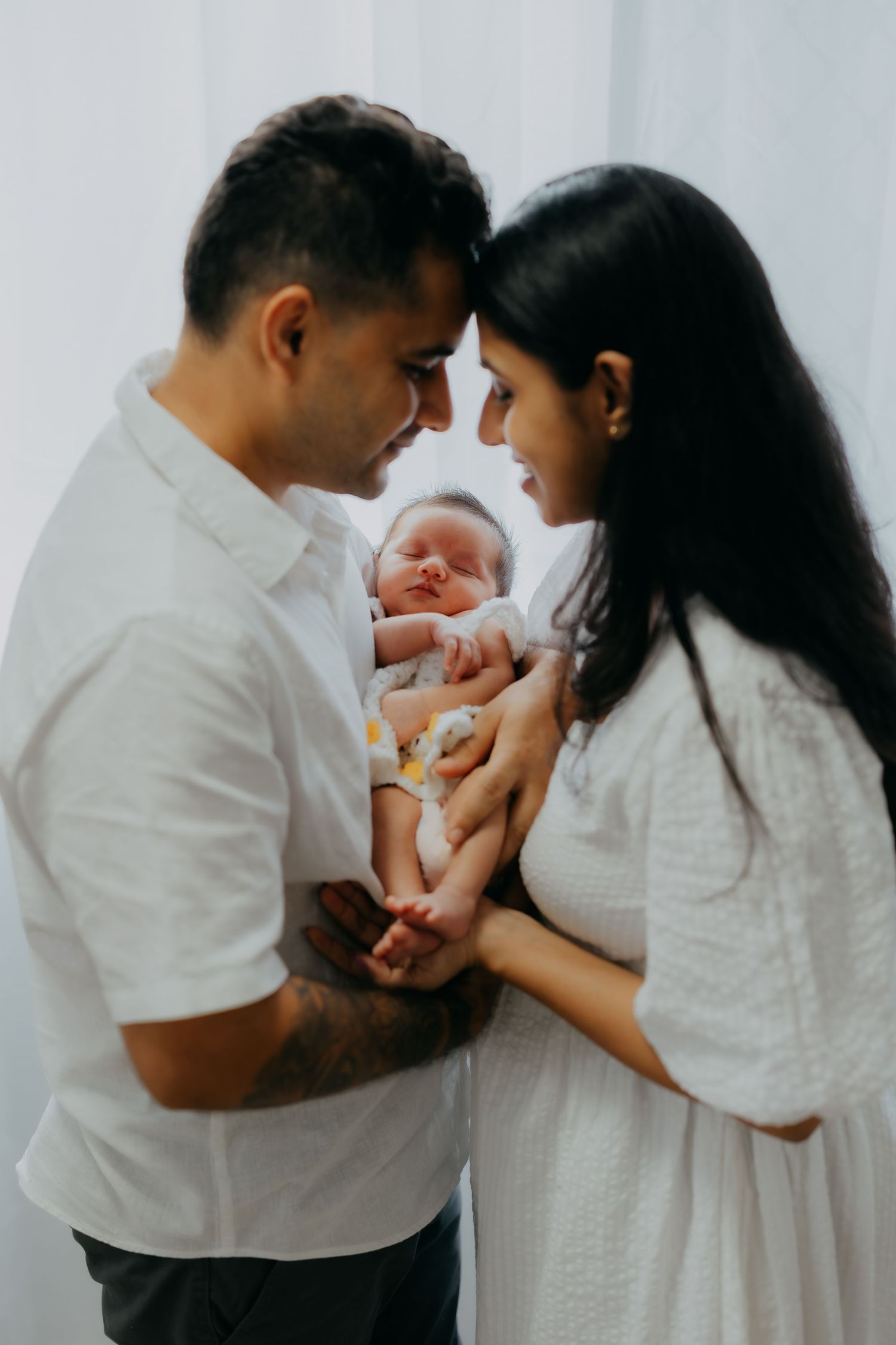 New parents in white outfits touch foreheads while gently holding their newborn baby together.