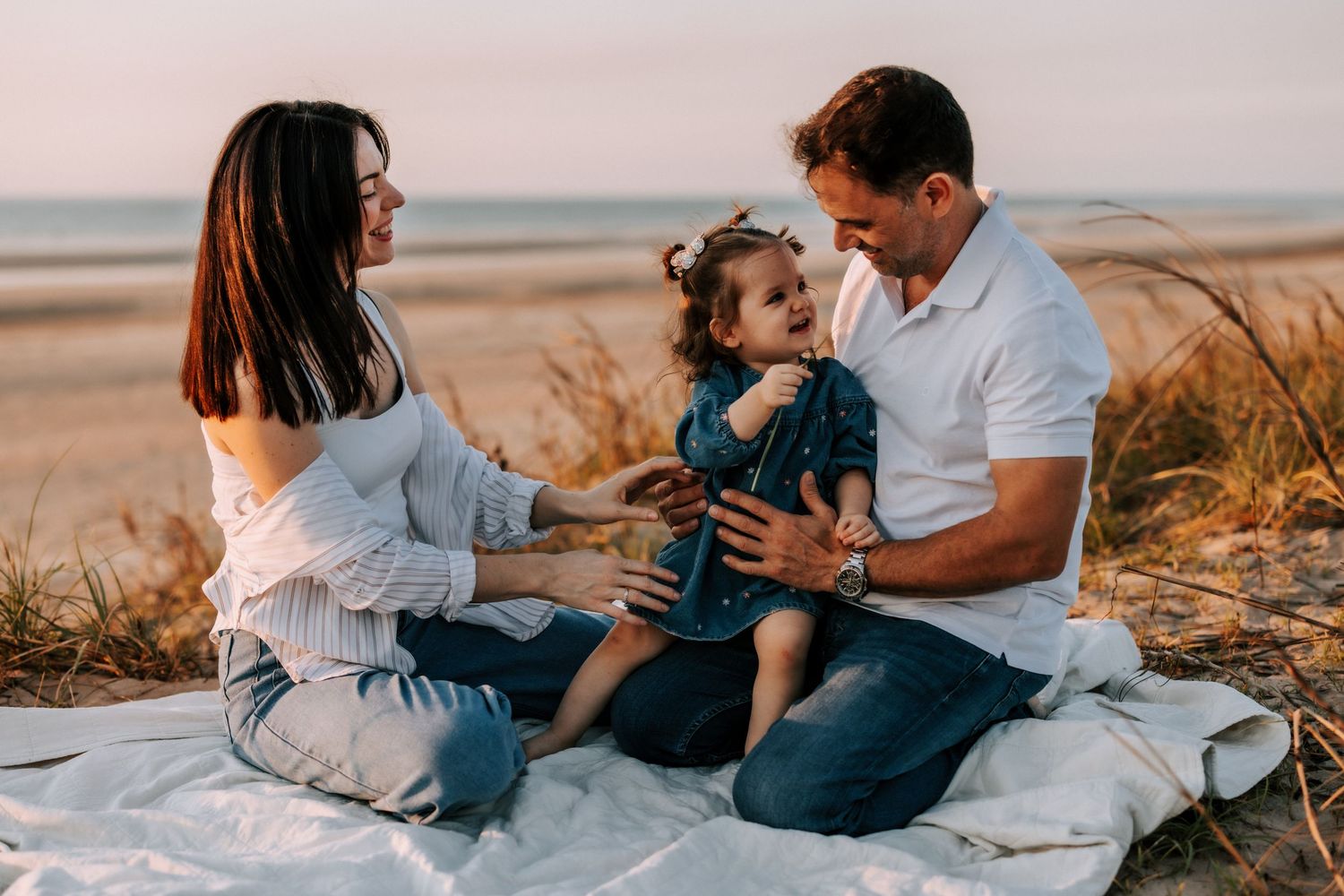 Happy couple sitting on a blanket in beach dunes, playing with their young toddler at sunset.
