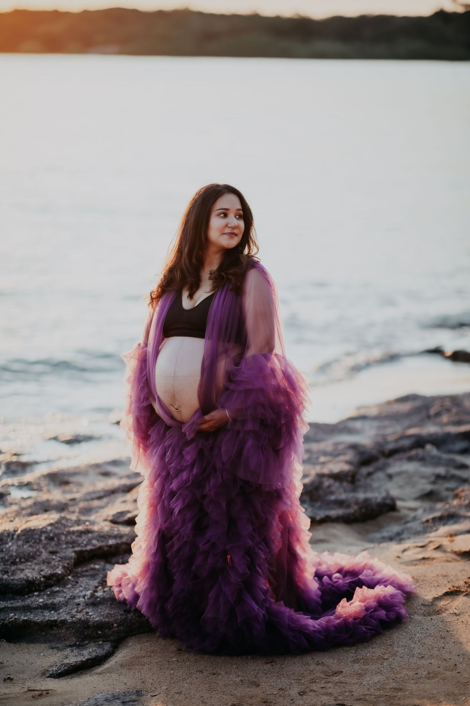 Pregnant woman in flowing purple tulle gown holding belly on rocky beach at sunset.