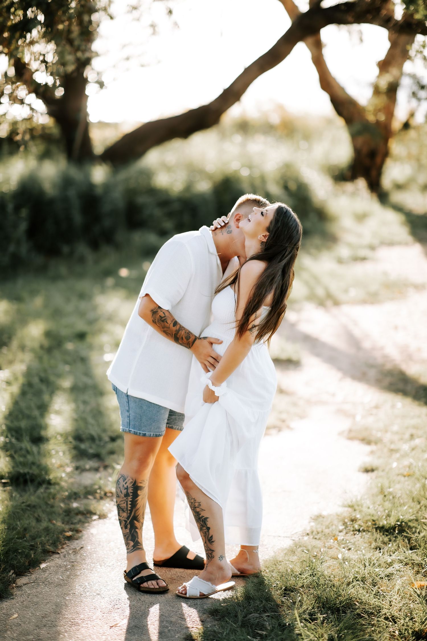 Tattooed couple kissing on a sunlit garden path, woman in white maternity dress.