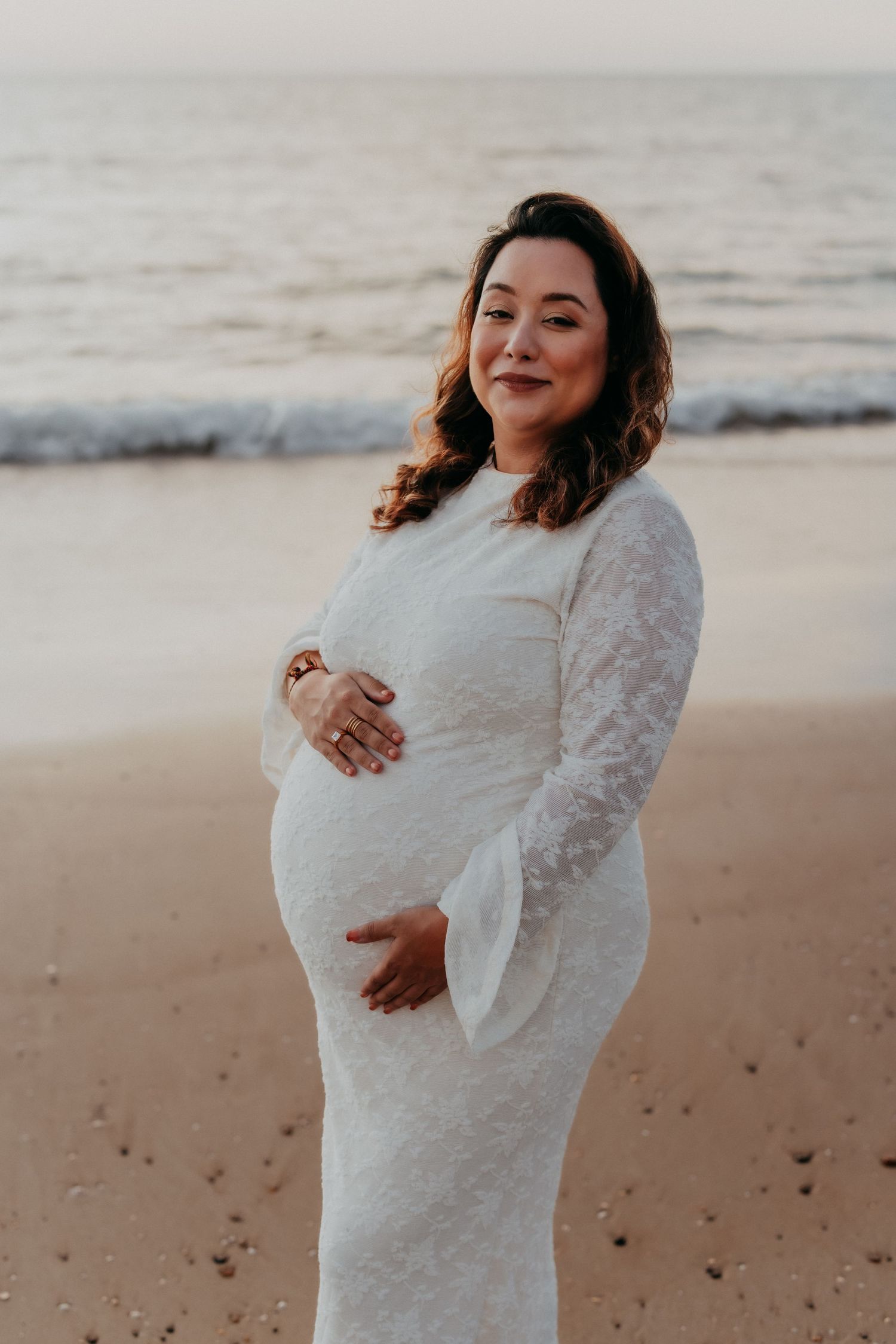 Smiling pregnant woman in white lace dress holding her belly on a sandy beach.