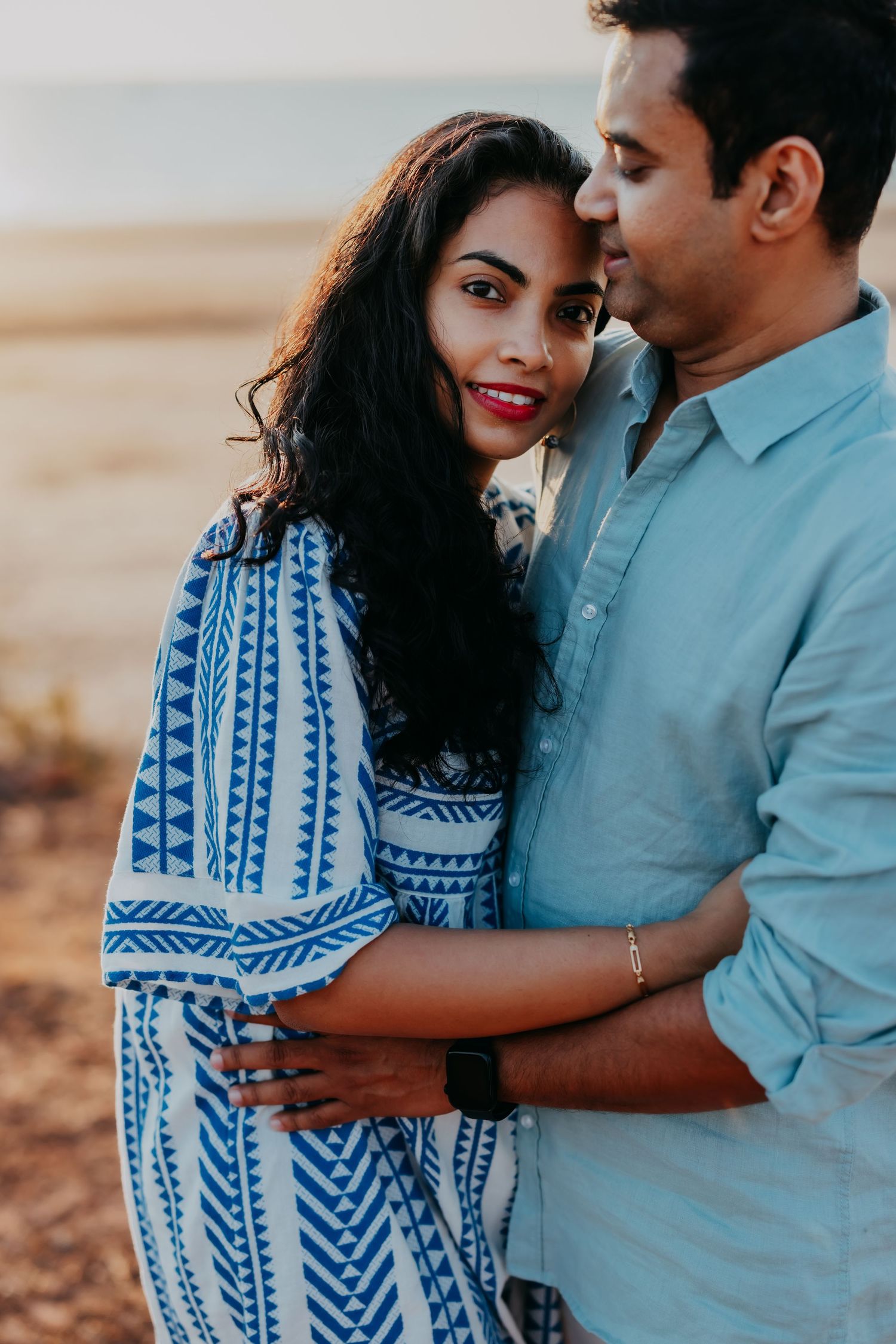 A loving couple embraces closely at sunset on a beach, the woman smiling at the camera.