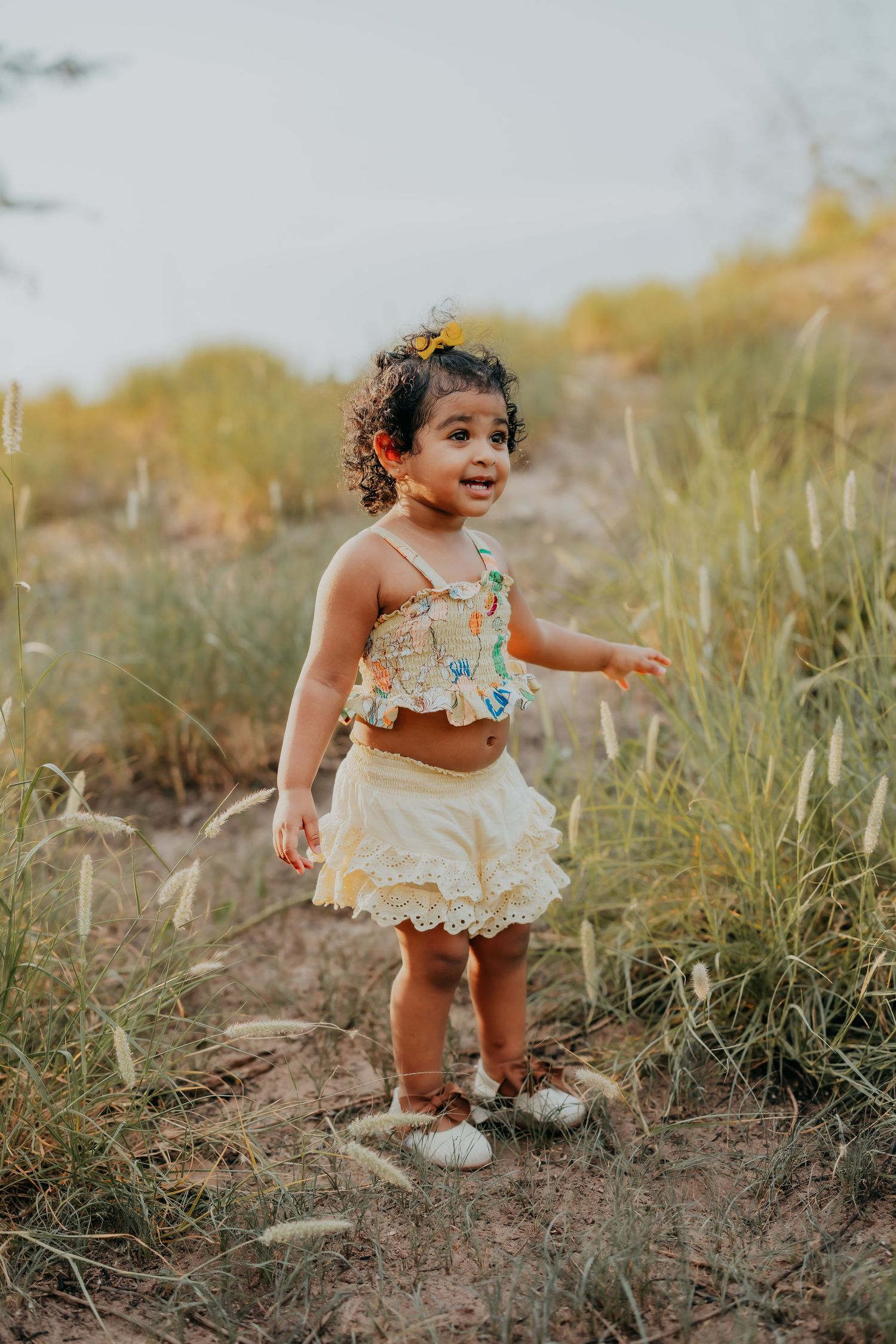 Adorable toddler girl in floral crop top and yellow skirt standing in tall grass during golden hour.