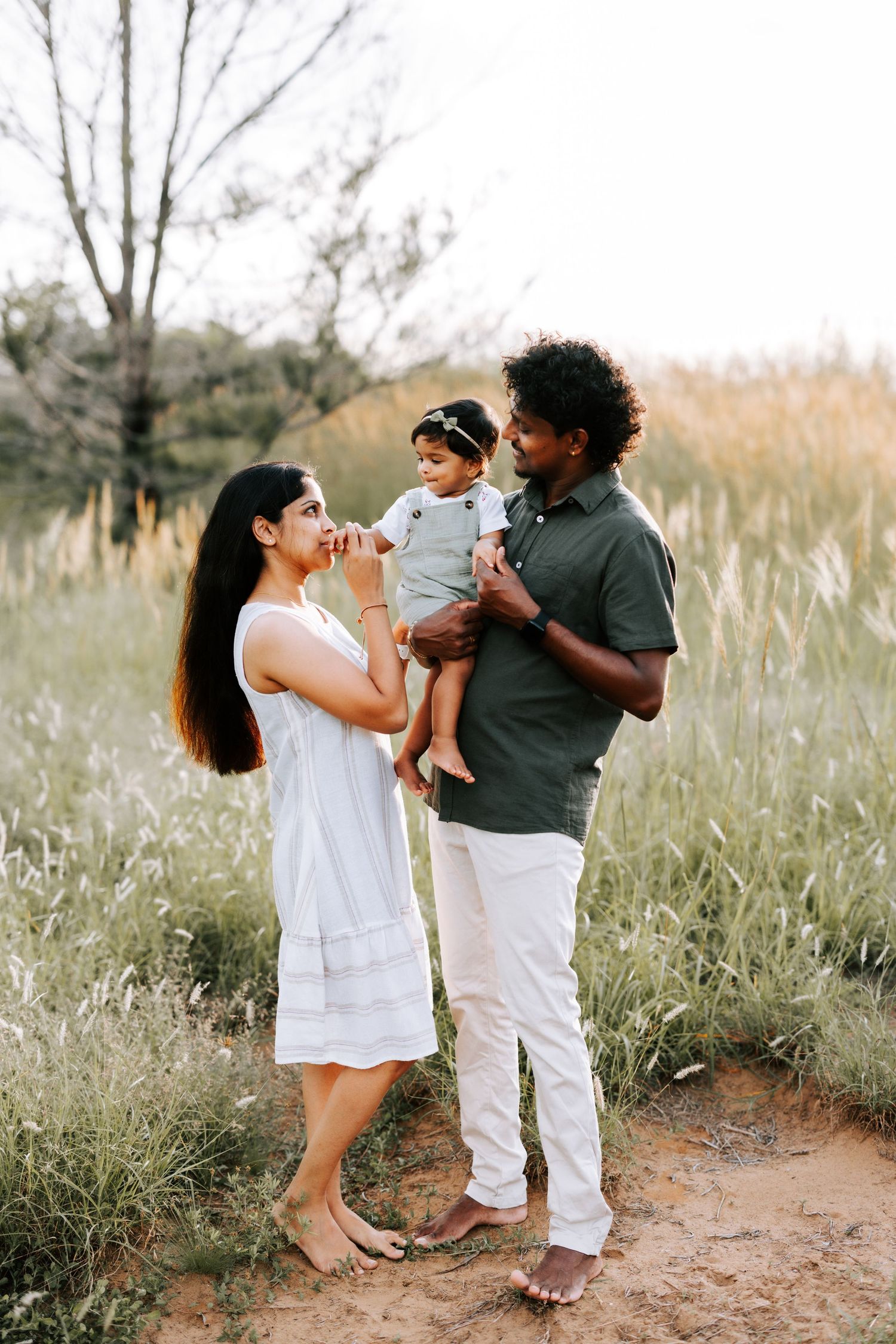 A couple stands in a golden grass field at sunset, lovingly holding their baby girl.