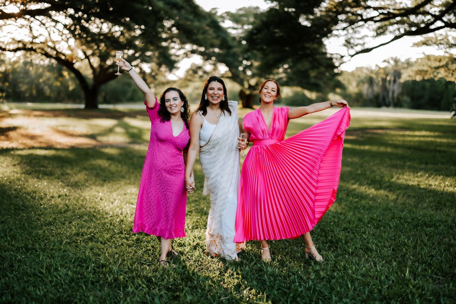 Three women in vibrant pink and white dresses celebrating outdoors in a lush green park.