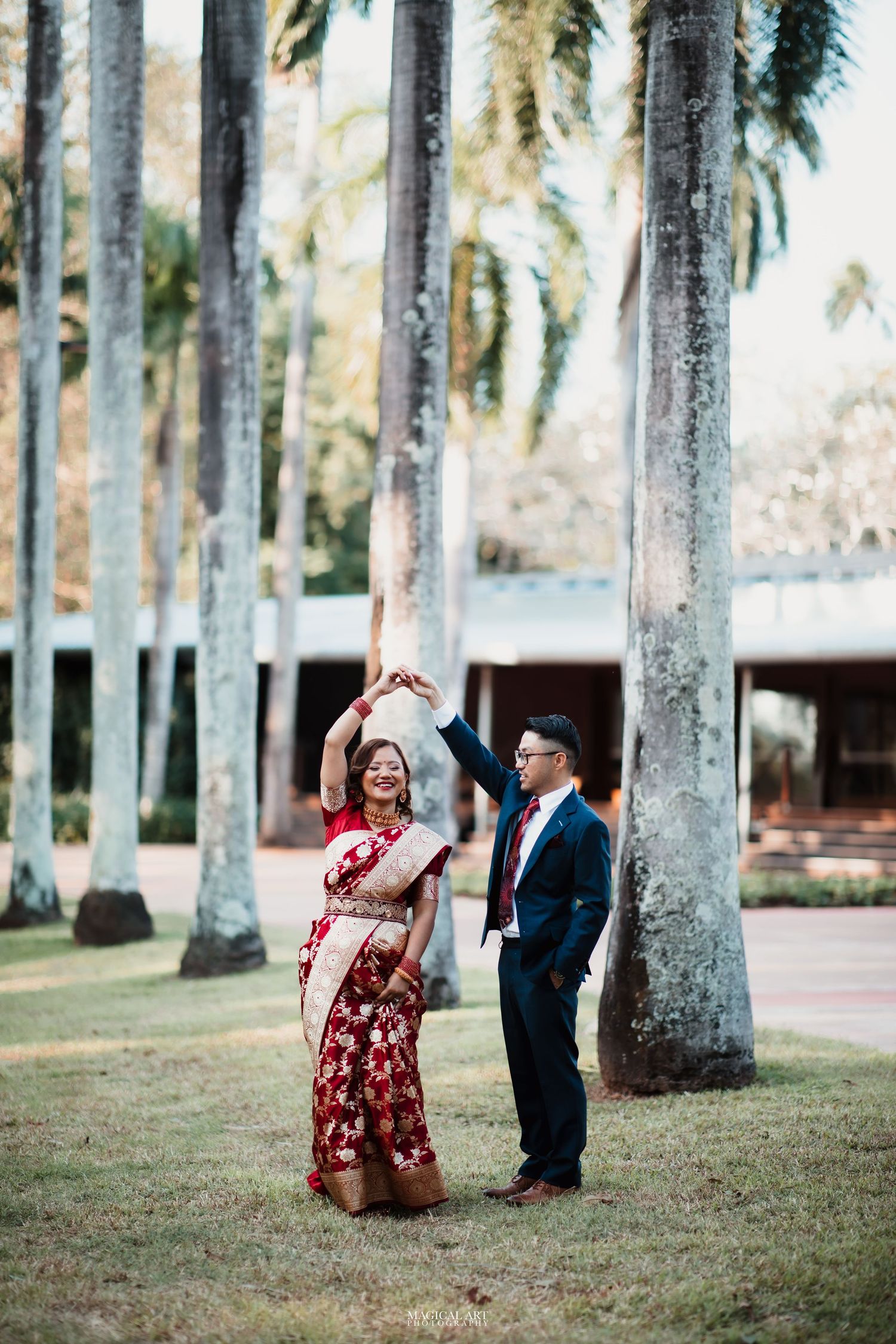 Groom in navy suit spinning bride in red floral saree among tall palm trees outdoors.