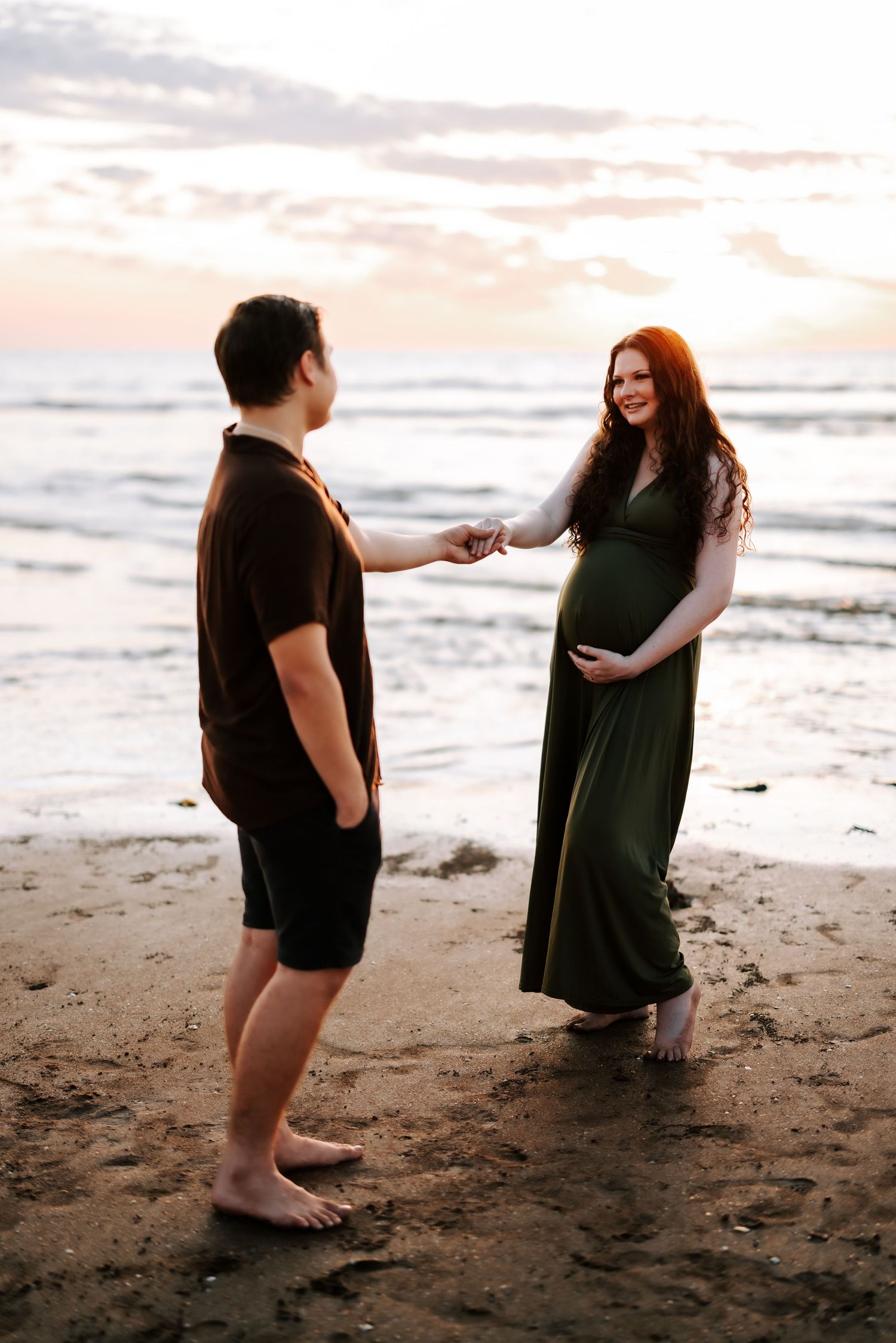 Expecting couple holding hands on a sandy beach at sunset, woman in green maternity gown.