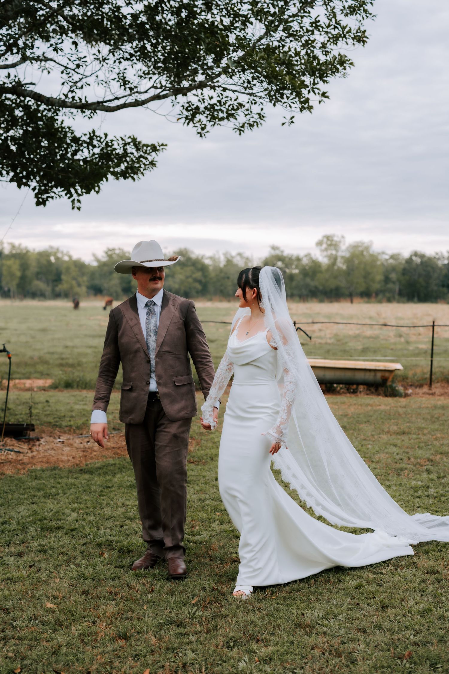 Bride and groom holding hands walking in a green field, wearing wedding dress with veil and brown suit with cowboy hat.