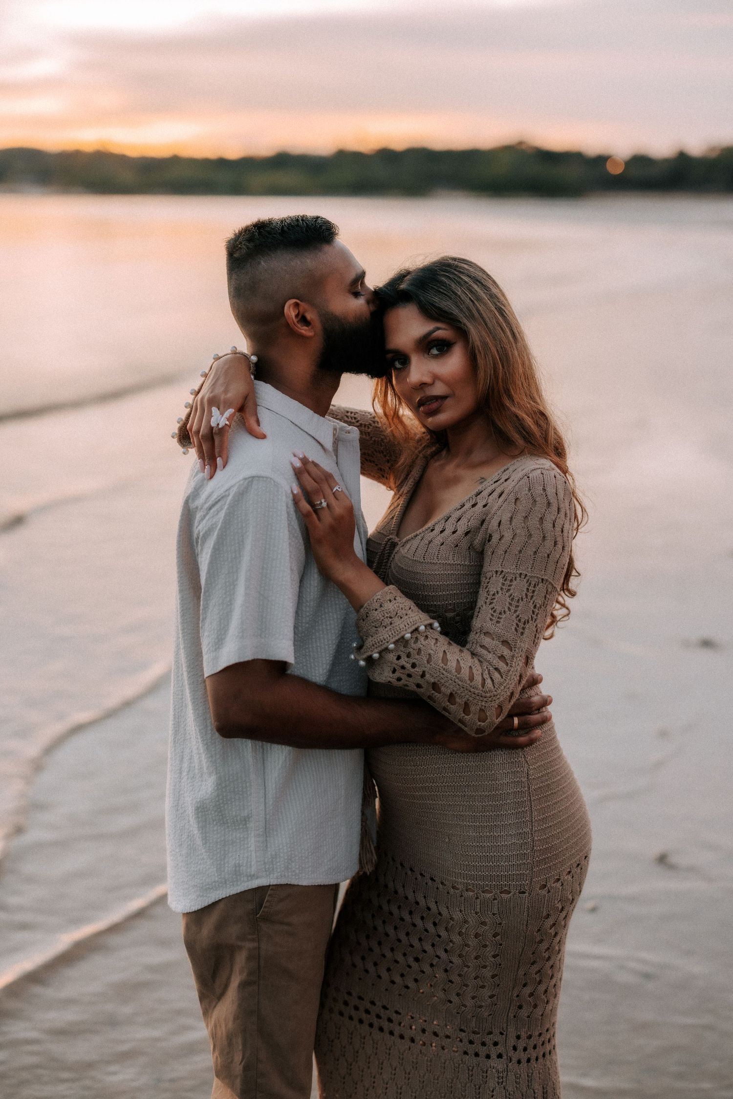 A couple embraces romantically at sunset on a beach, woman in knit dress, man in white shirt kissing her forehead.