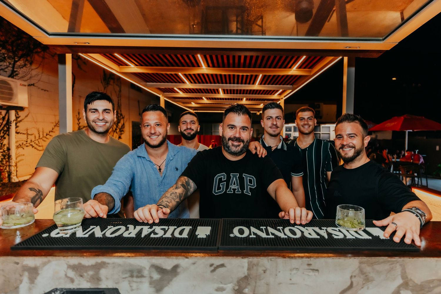 Group of seven smiling men posing together at an outdoor bar with Disaronno branding on the counter.