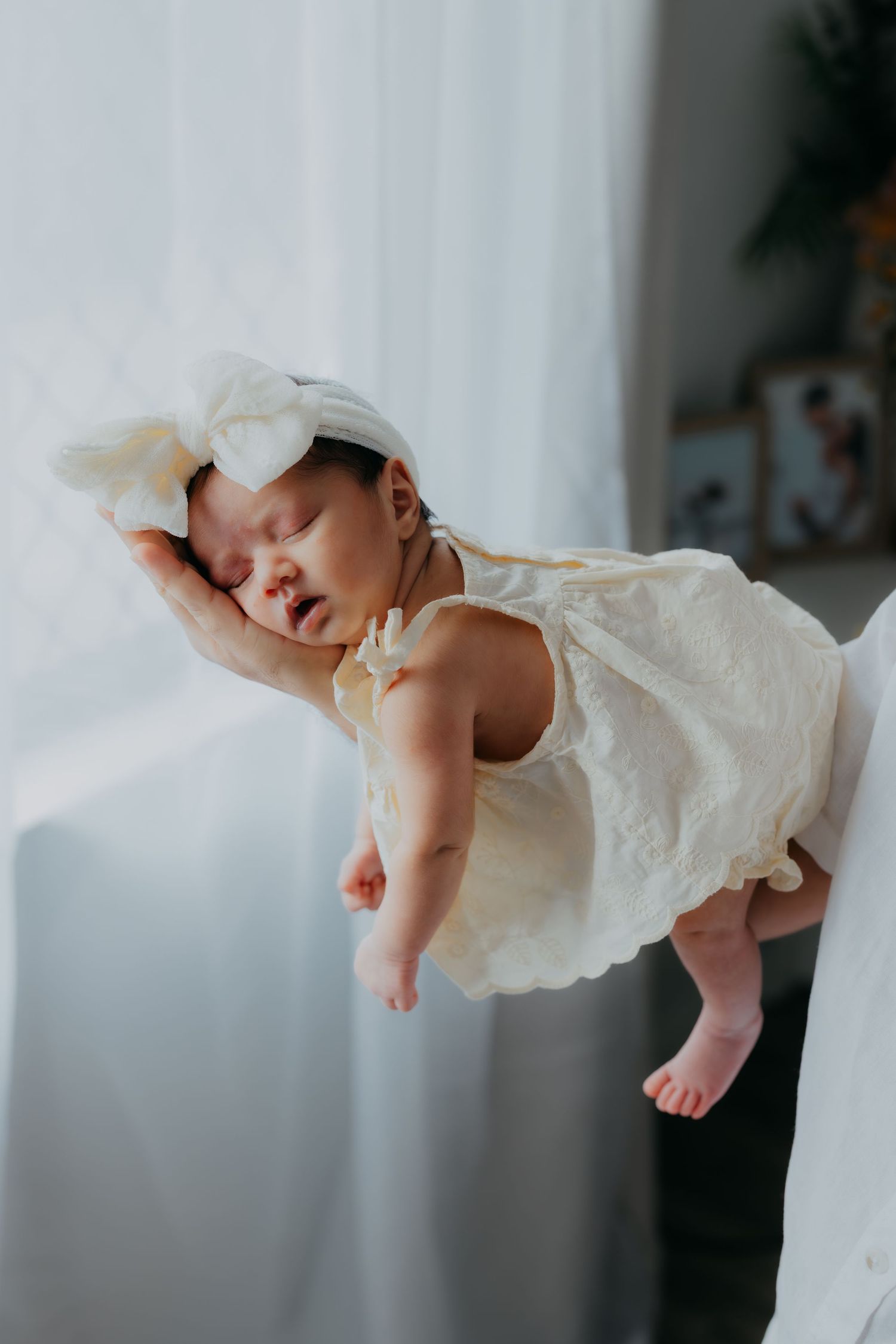 Sleeping newborn baby girl in cream dress and bow headband being held near a window.