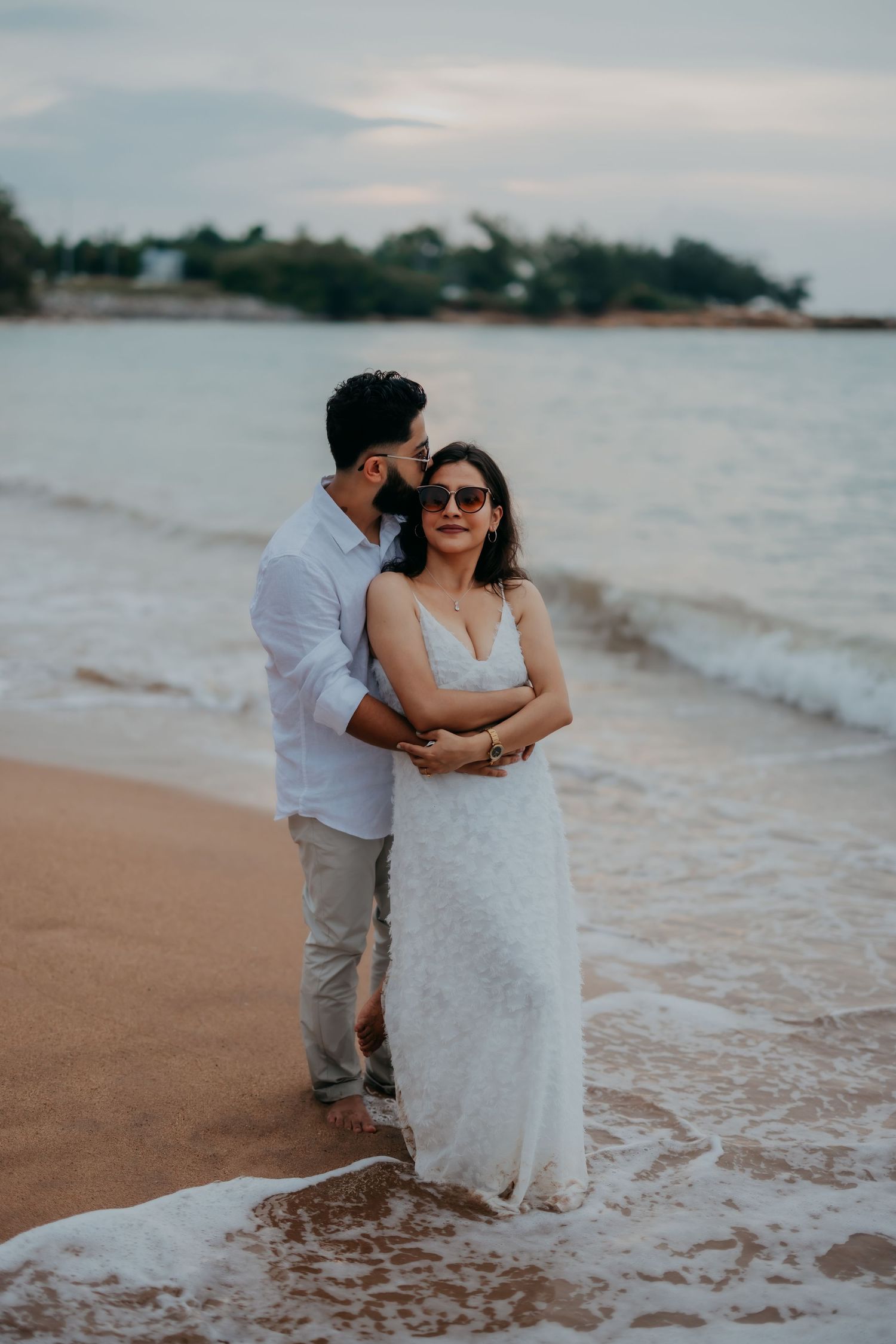 Groom embracing bride from behind on sandy beach at dusk, both in white attire with ocean waves.