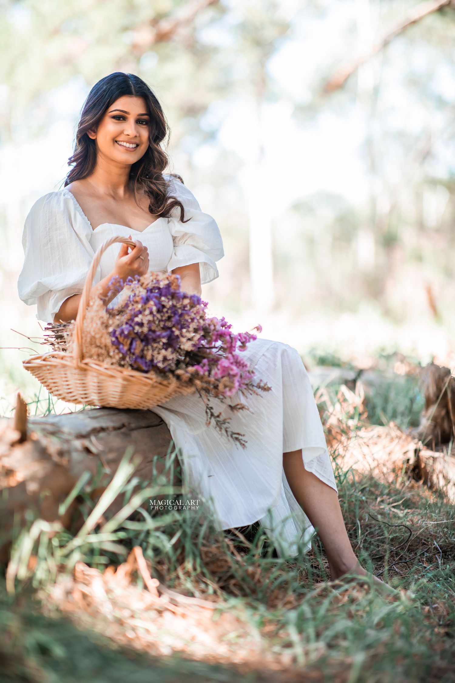 Smiling woman in white dress holds flower basket while sitting outdoors in sunlit forest setting.