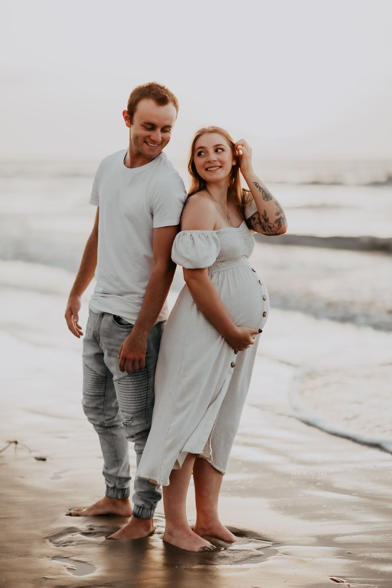 Smiling couple standing barefoot on beach, pregnant woman in white dress.