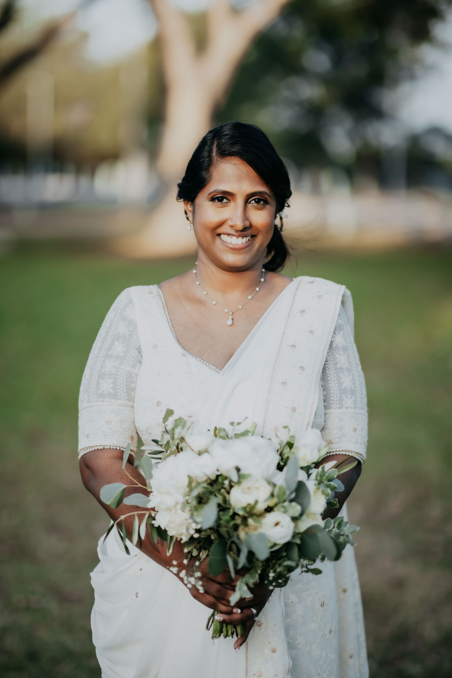 Smiling South Asian bride in white embroidered saree holds white and green bouquet in outdoor park.