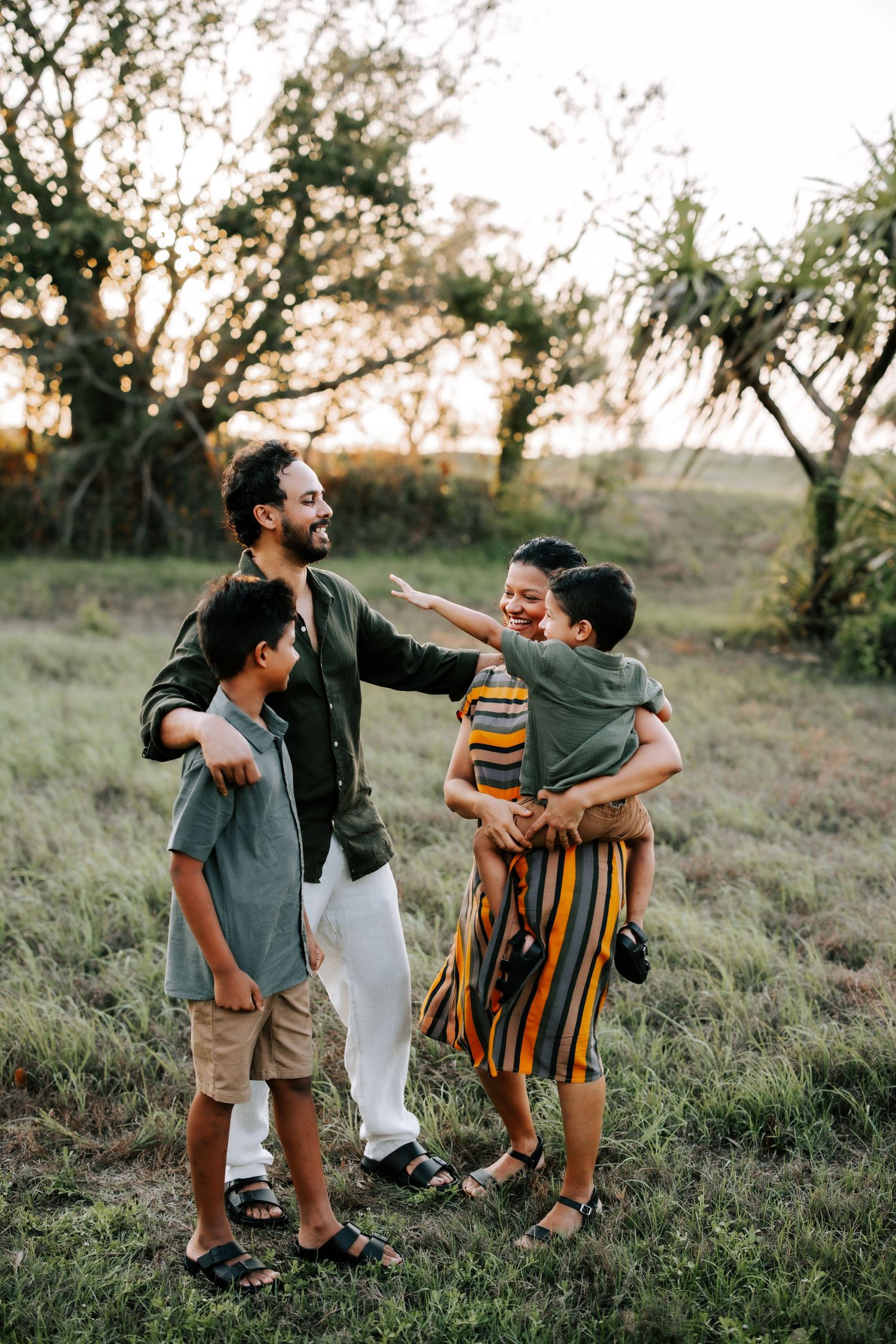 A joyful family of four laughs together in a golden sunset field, with parents and two boys enjoying outdoor time.