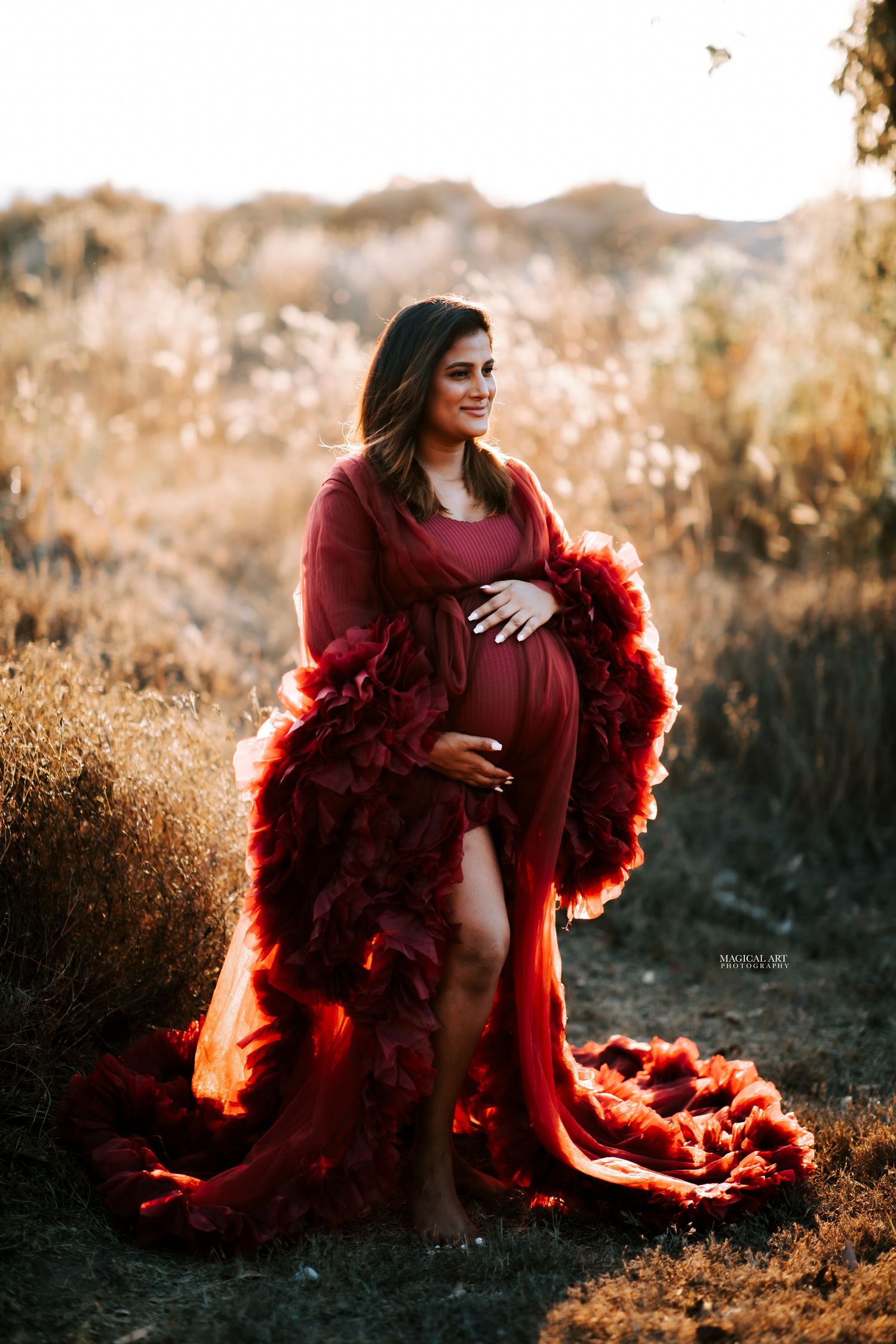 Pregnant woman in elegant dark red ruffled maternity gown posing outdoors at golden hour.