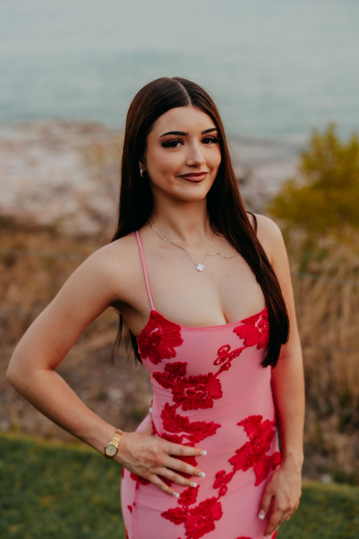 Young woman in pink floral dress posing outdoors near waterfront at golden hour.