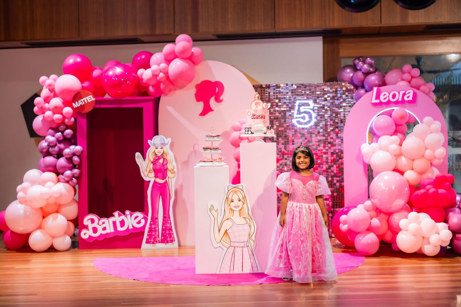 Young girl in pink dress posing at a Barbie themed 5th birthday party with balloon arch and character cutouts.
