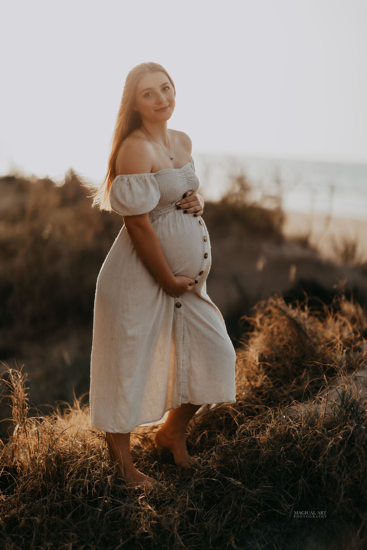 Pregnant woman in white off-shoulder dress holding belly in golden sunset field.