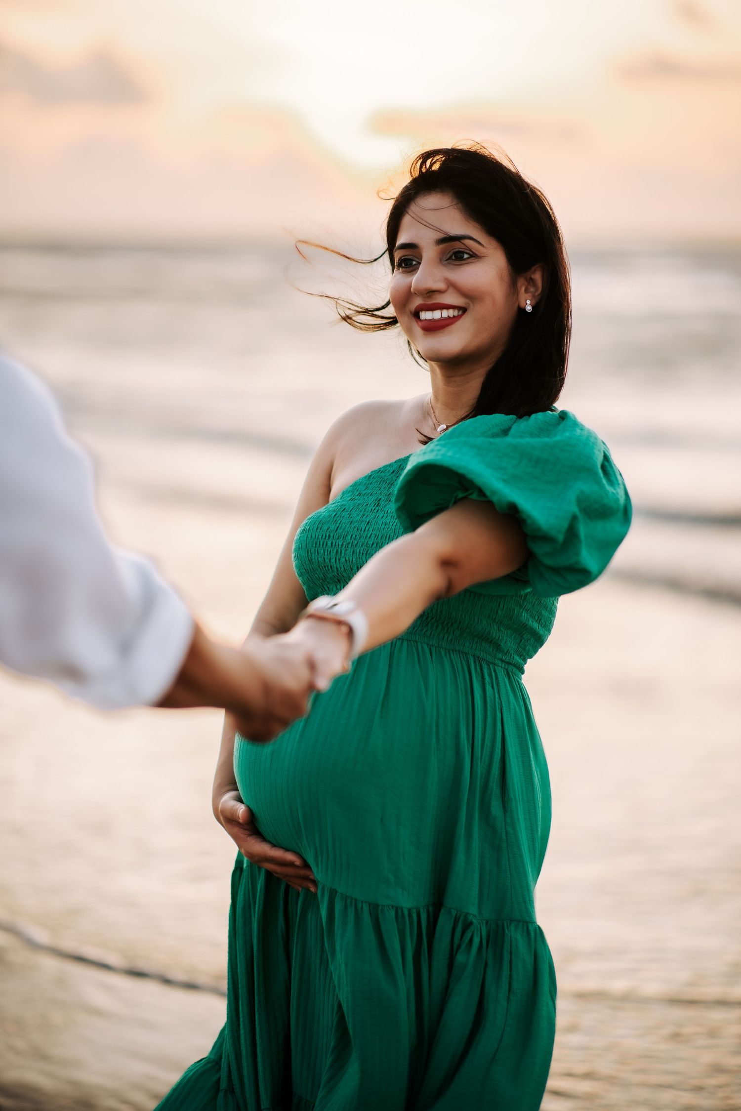 Pregnant woman in green dress holds partner's hand on beach at sunset.