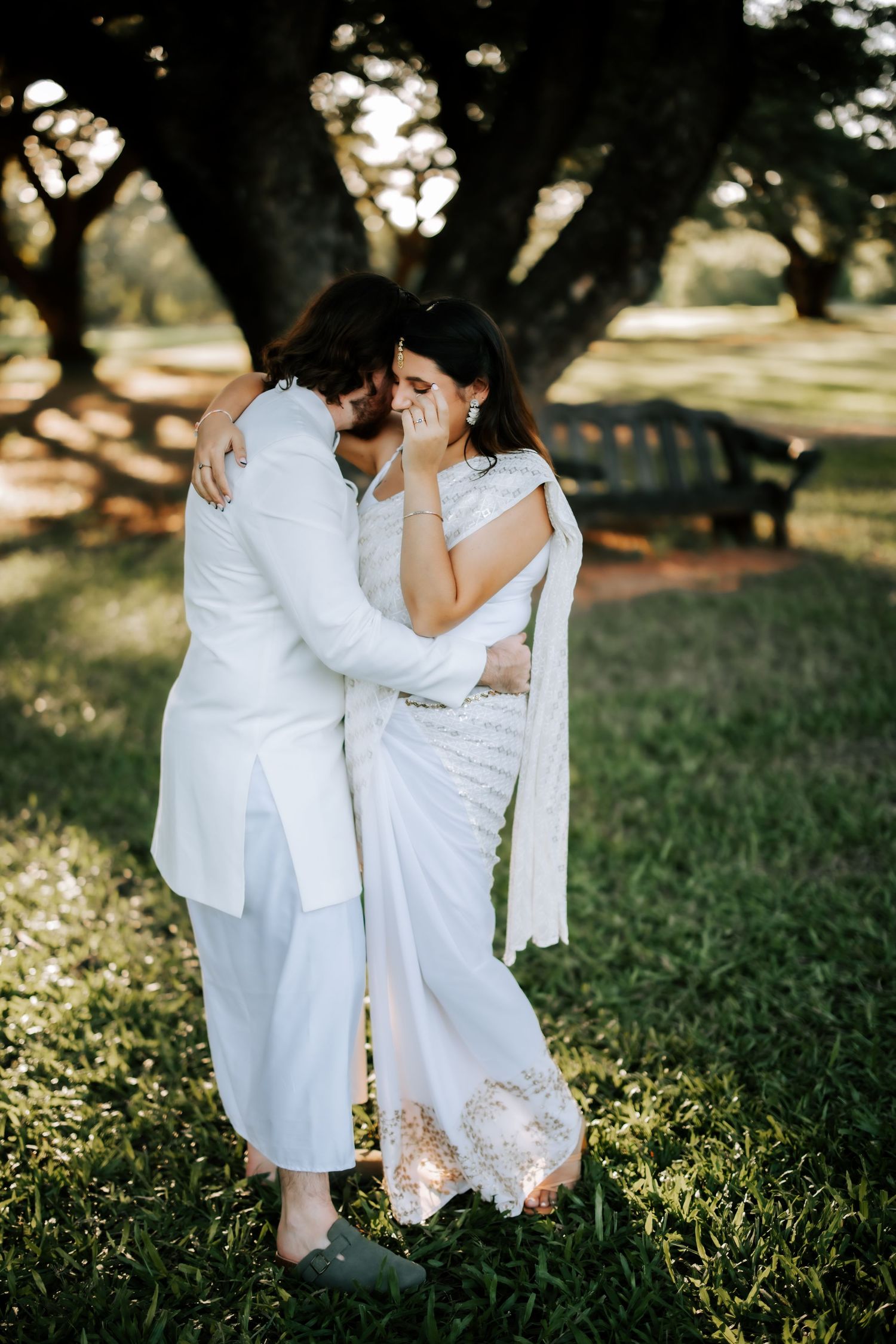Emotional bride in white saree embraces groom under large trees during golden hour wedding portraits.