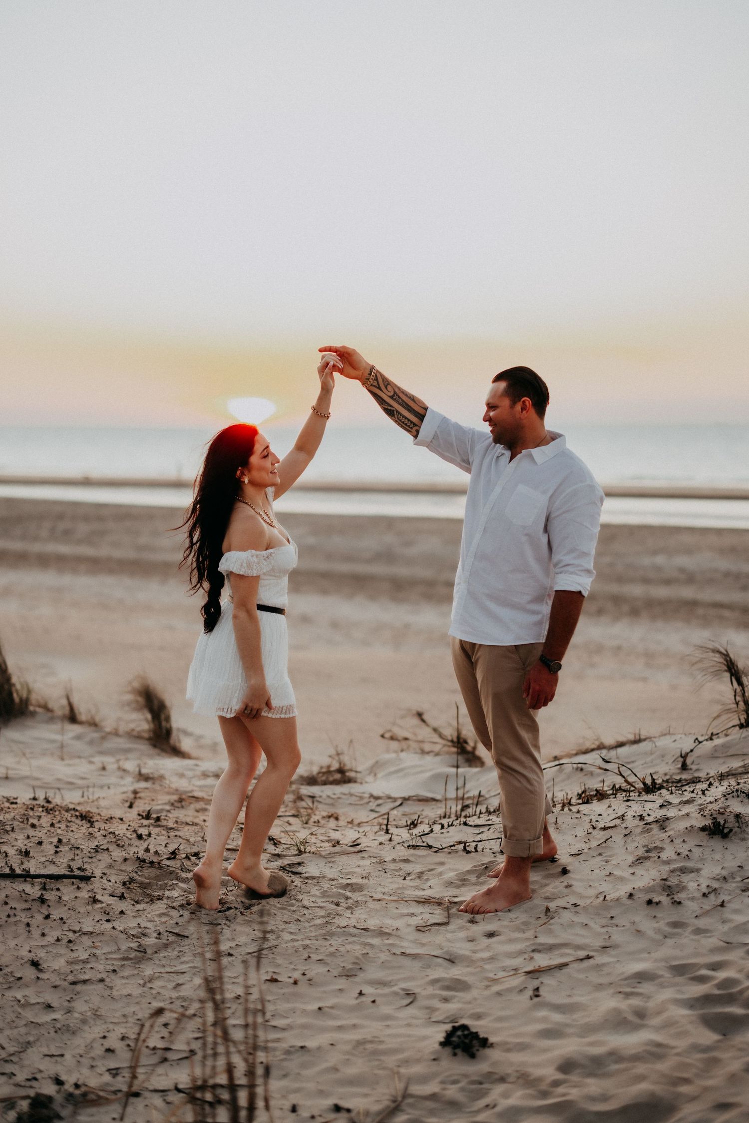 Couple dancing barefoot on sandy beach at sunset, man spinning woman in white dress near ocean shore.