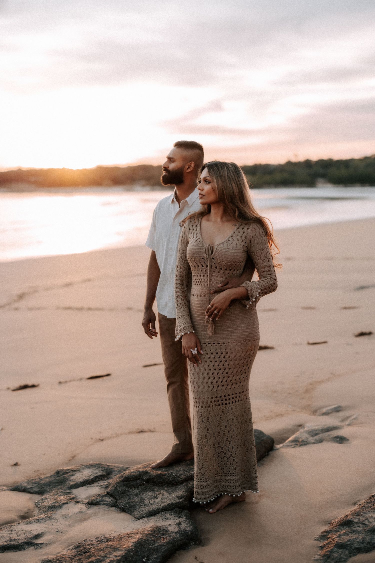 Couple standing on a rocky beach at sunset, woman in crochet maxi dress, man in white shirt and tan pants.