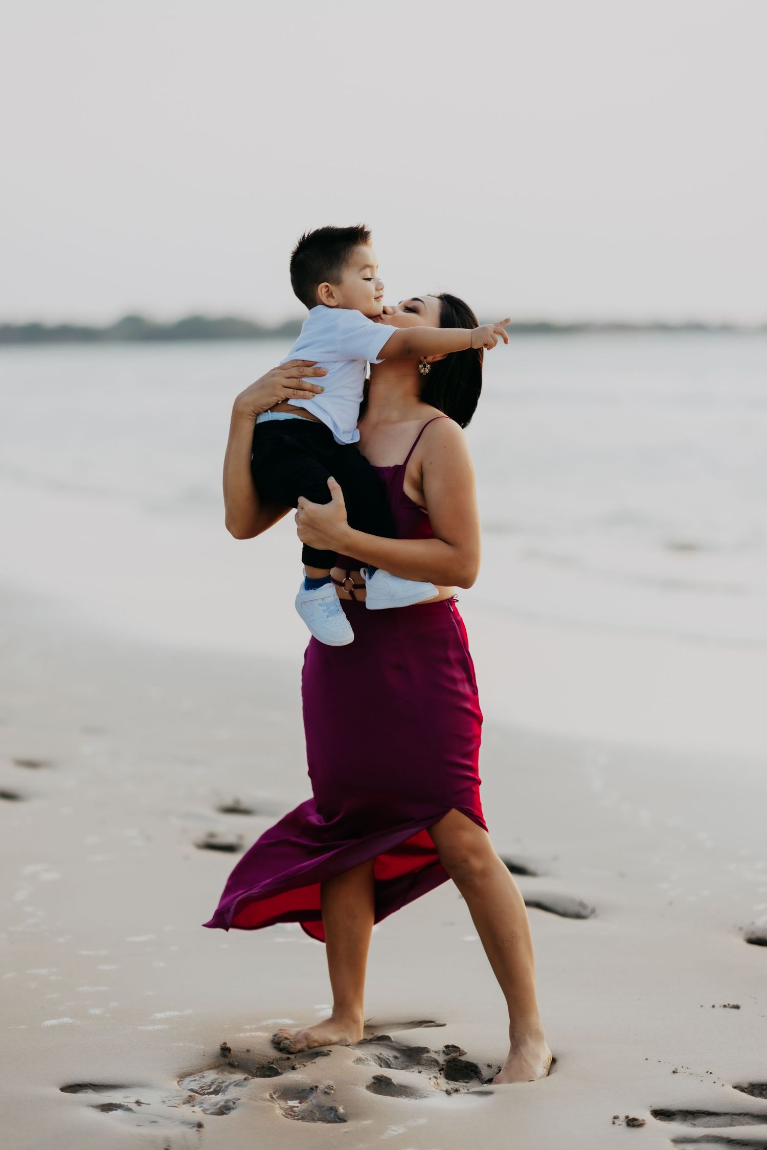A mother in a purple dress lovingly kisses her young son while walking on a sandy beach.