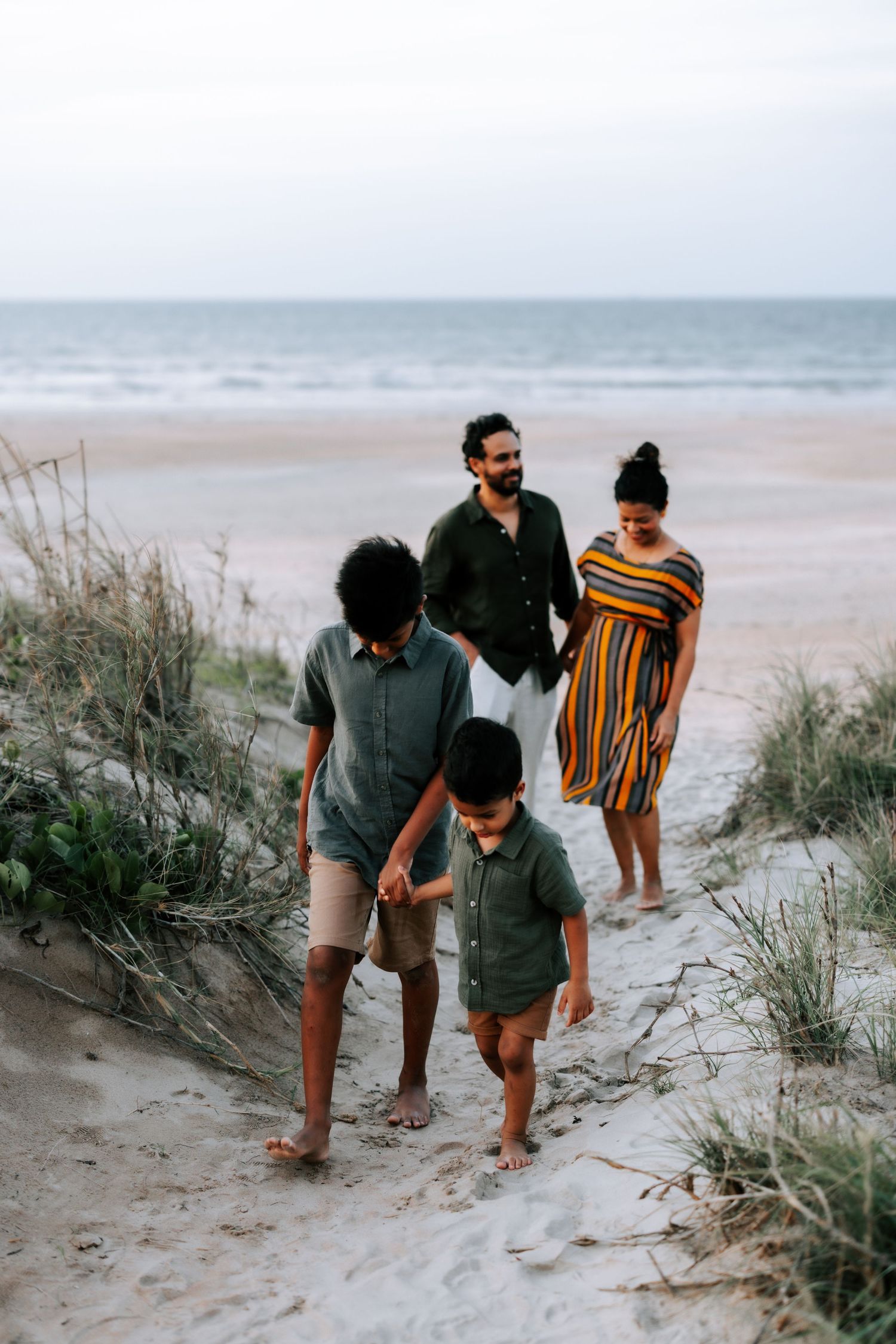 A happy family of four walks along sandy beach dunes near the ocean at twilight.