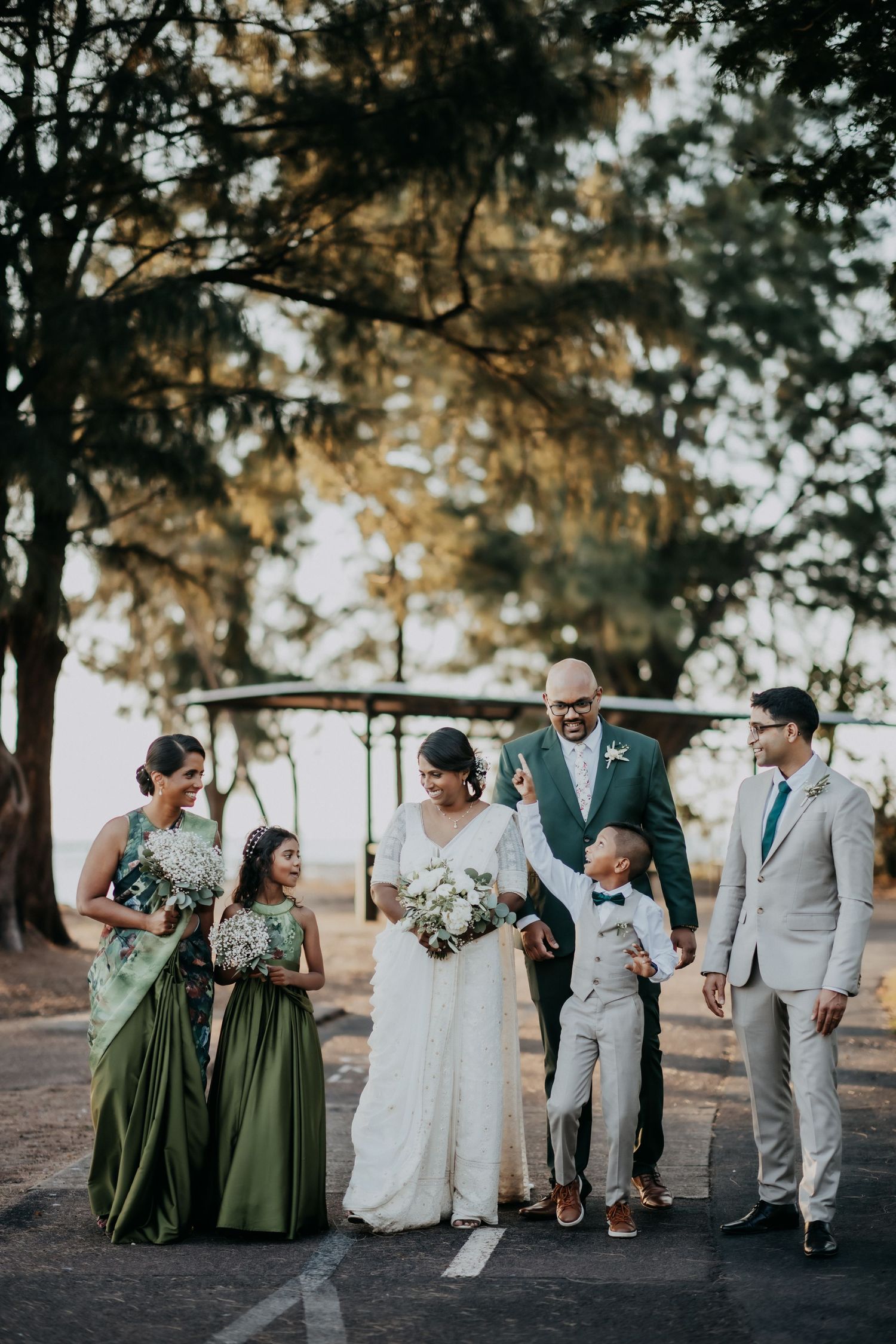 Wedding party poses outdoors in green bridesmaid dresses and grey suits with bride in white saree.
