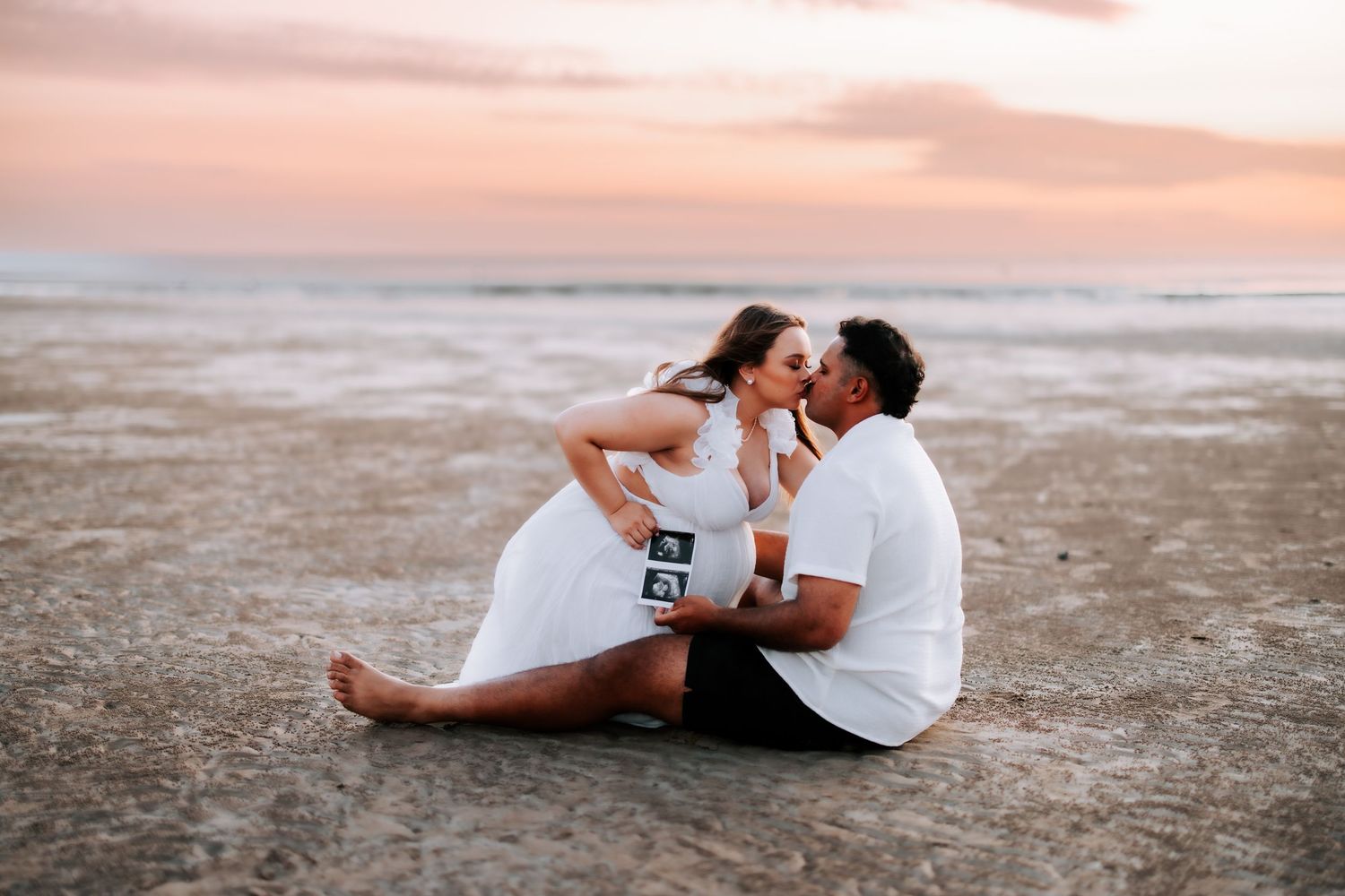 Couple sitting on beach at sunset sharing a kiss while holding ultrasound photos on the sand.