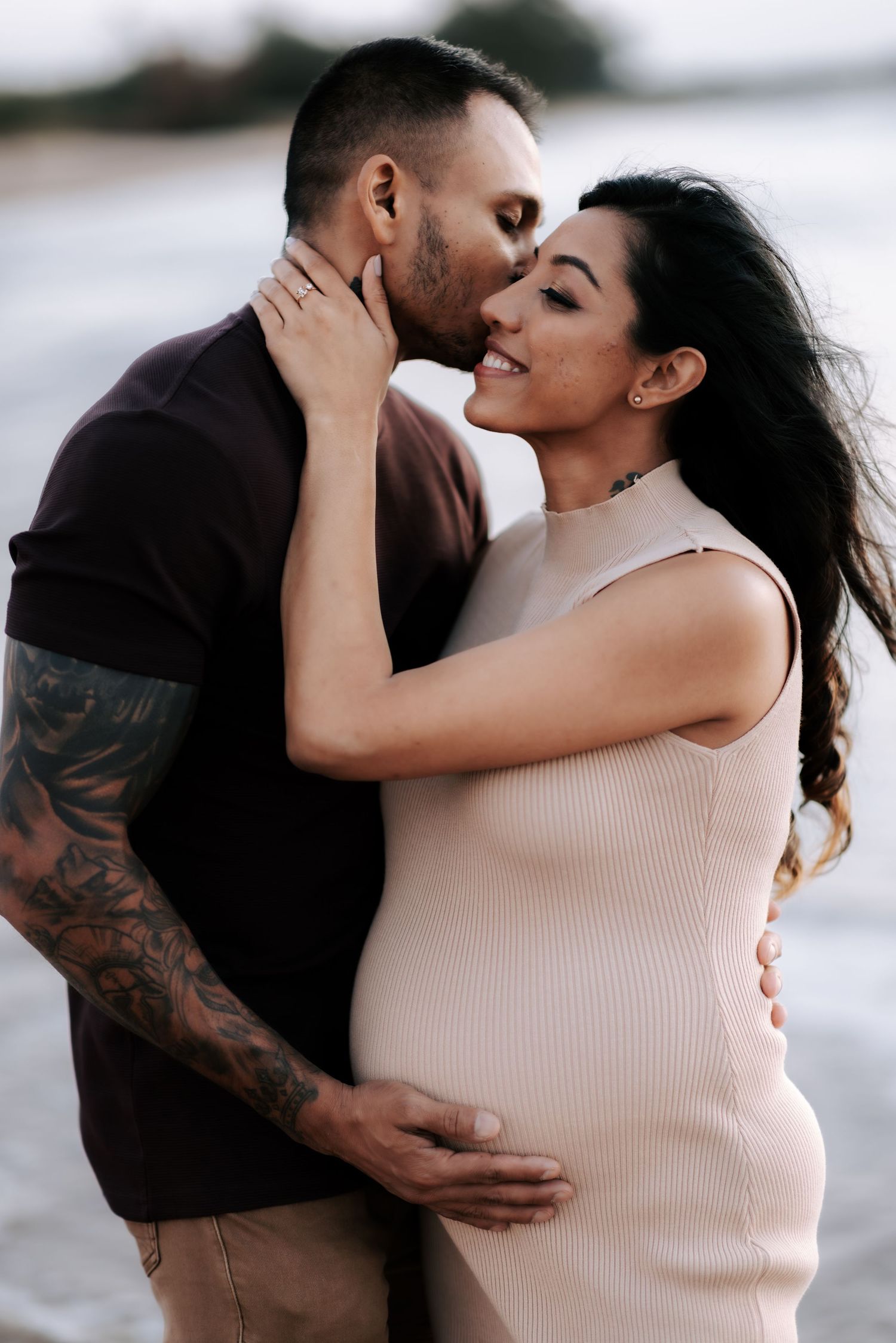 Tattooed man kisses smiling pregnant woman in beige dress on breezy beach at sunset.